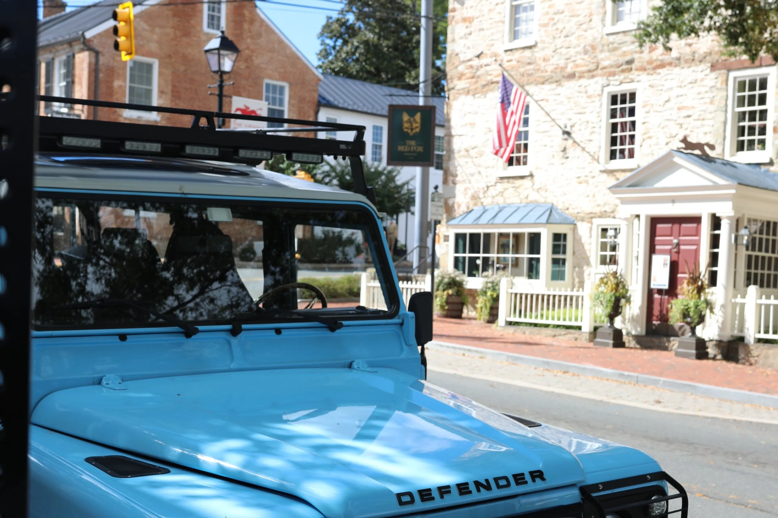 Front view of a light blue Land Rover Defender parked on a city street with brick buildings and a restaurant sign in the background.