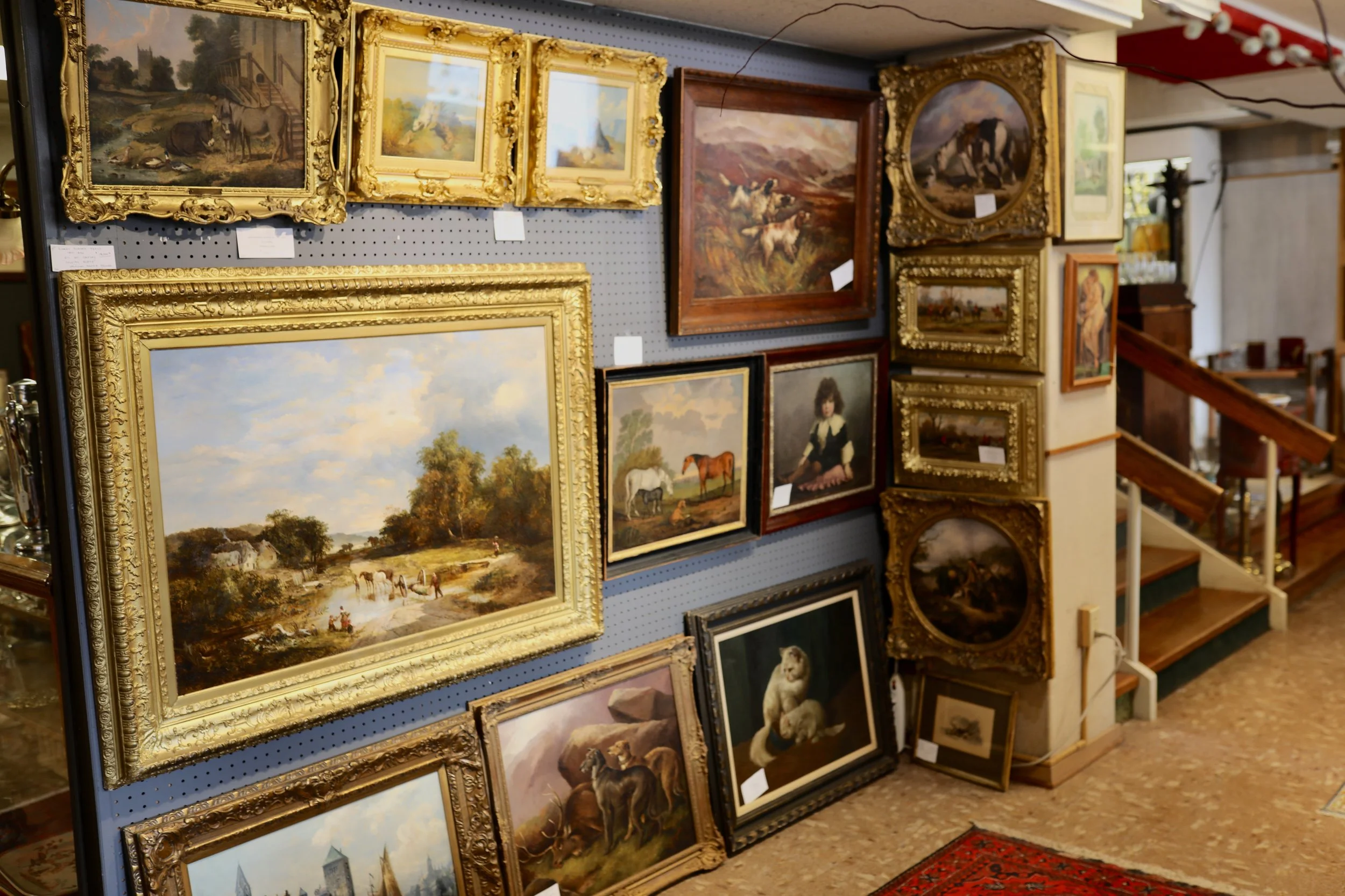 A wall filled with framed paintings, mostly landscapes and animal scenes, displayed on a gray pegboard in an antique shop. There are various sizes of ornate and simple frames, with some leaning against the wall and others hung. The floor features a red patterned rug near the bottom right corner.
