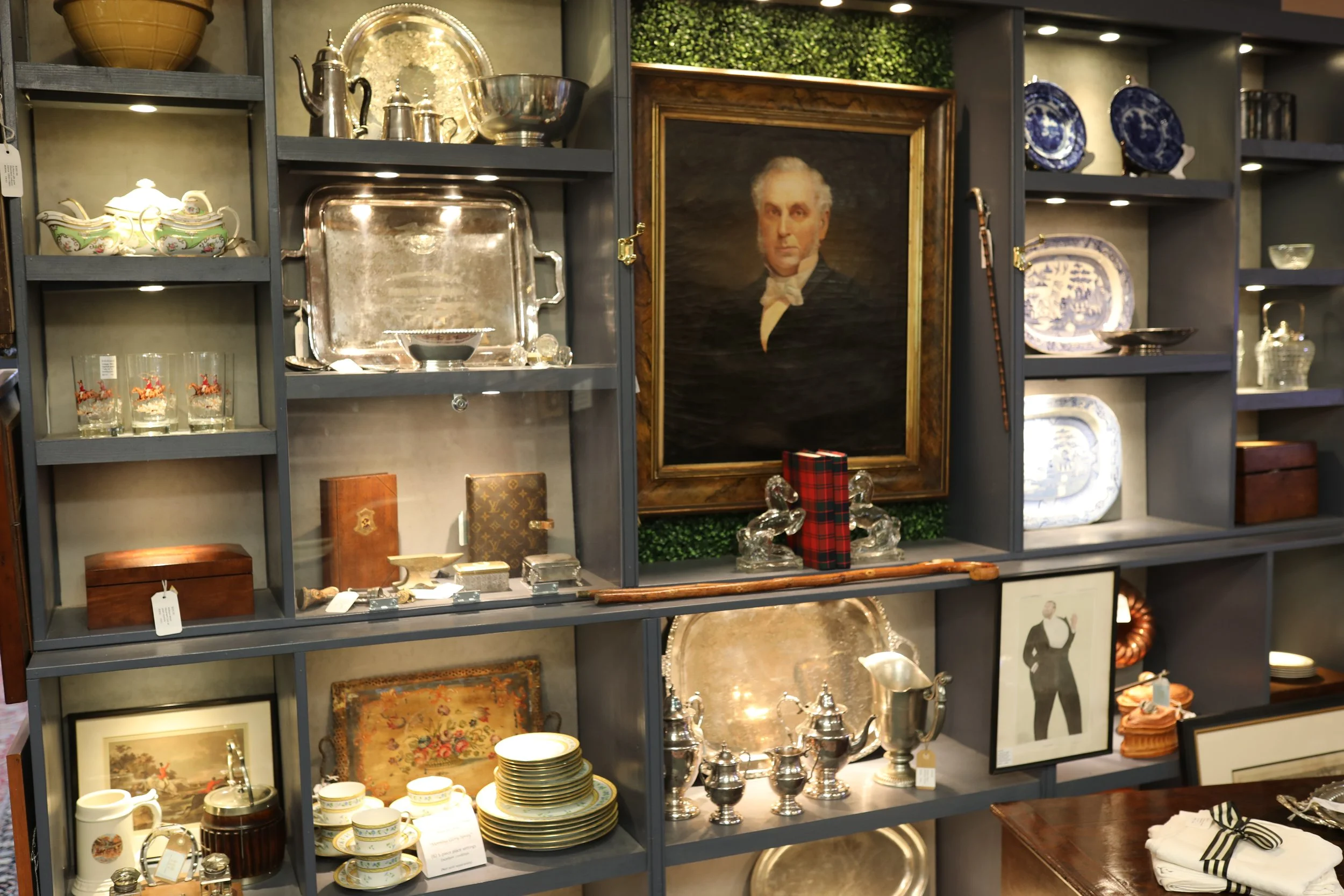 A display shelf filled with antique silverware, china dishware, framed portraits, and decorative items in an antique shop.