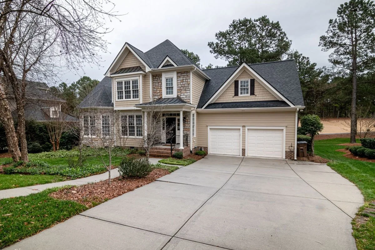 A two-story suburban house with beige siding, stone accents, and black shutters. There is a concrete driveway leading to a double garage and a small front porch with steps. The house is surrounded by a yard with grass, trees, and shrubs.