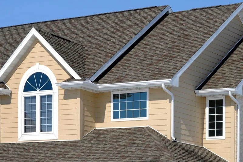 Close-up of the upper part of a beige house with three white-framed windows, brown shingle roof, and white gutters, against a clear blue sky.