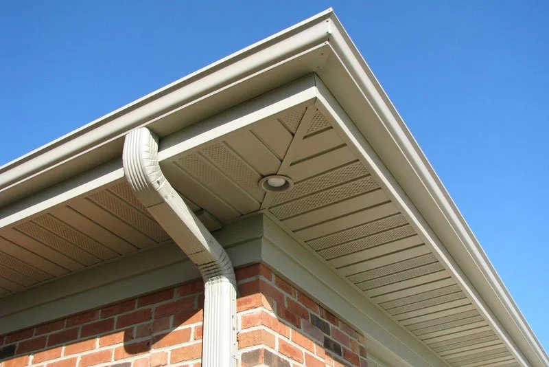 Close-up of the corner of a brick house roof showing white soffit, fascia, gutter, and a round vent, with a clear blue sky in the background.