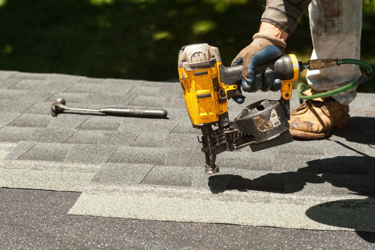 A worker in gloves installs asphalt shingles on a roof using a nail gun.