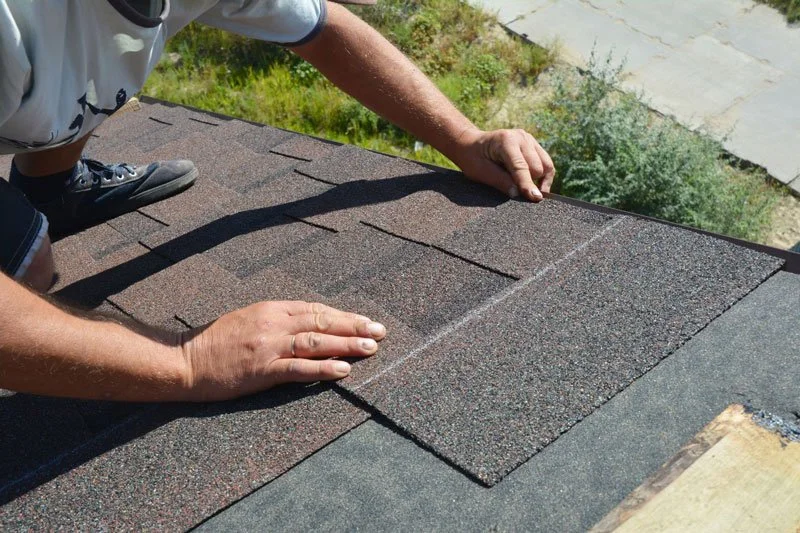 A person installing asphalt shingles on a roof, with their hands smoothing a shingle in place.