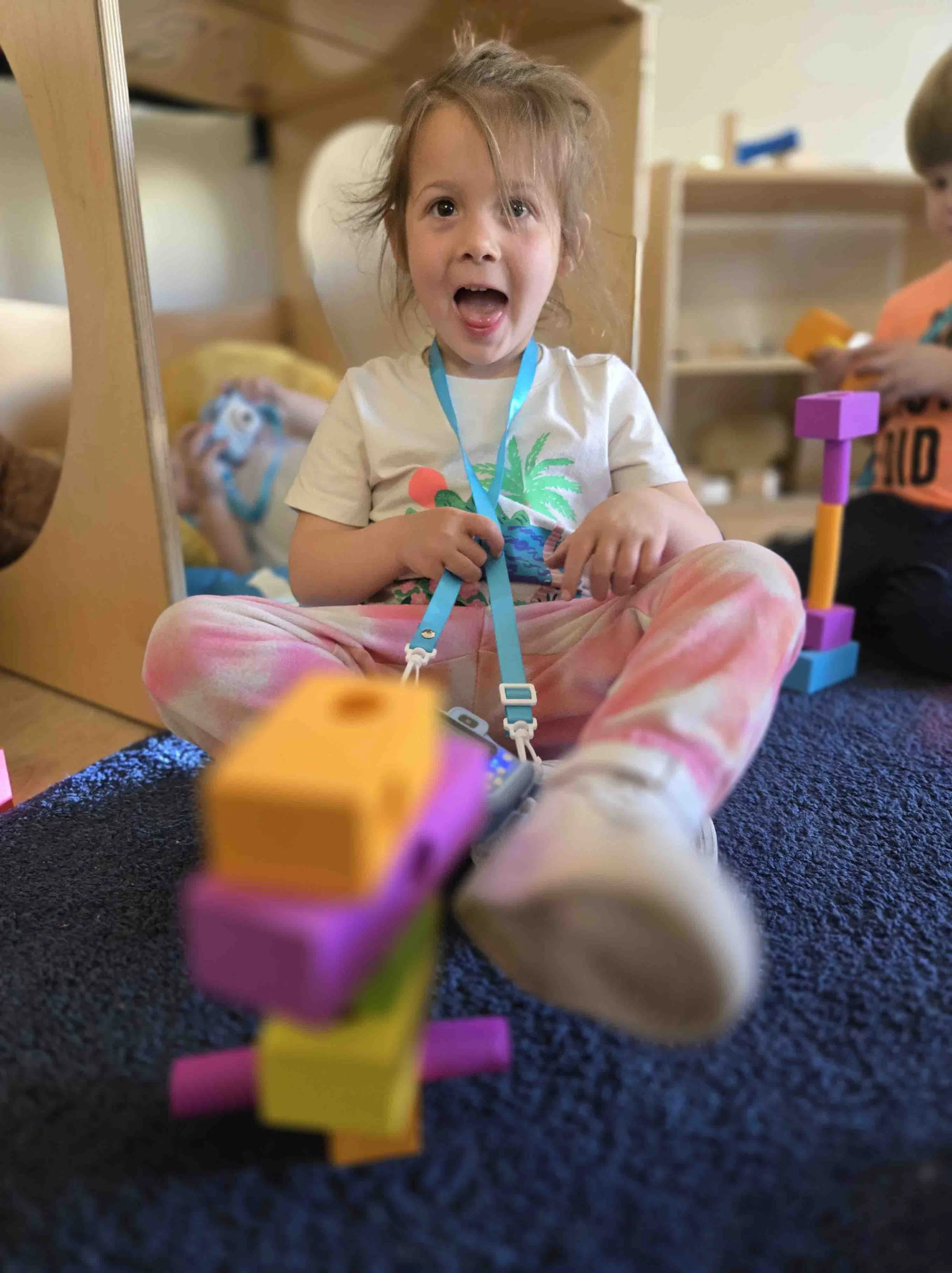 A young girl sitting on a dark carpet, smiling and holding a toy with colorful blocks and a string. There is a mirror behind her and other children playing in the background.