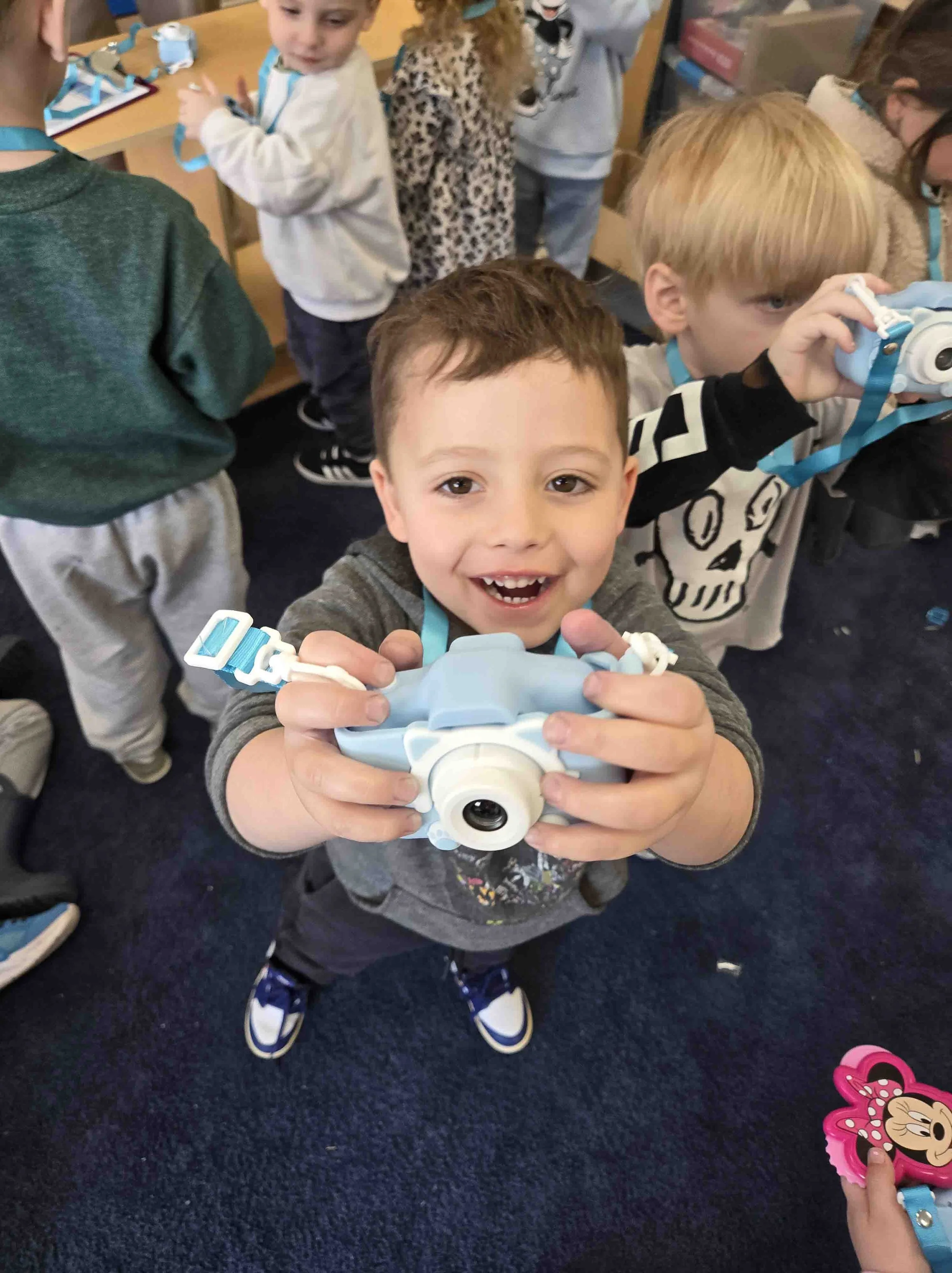 A group of children in a classroom holding and examining toys, with a boy smiling at the camera in the foreground.