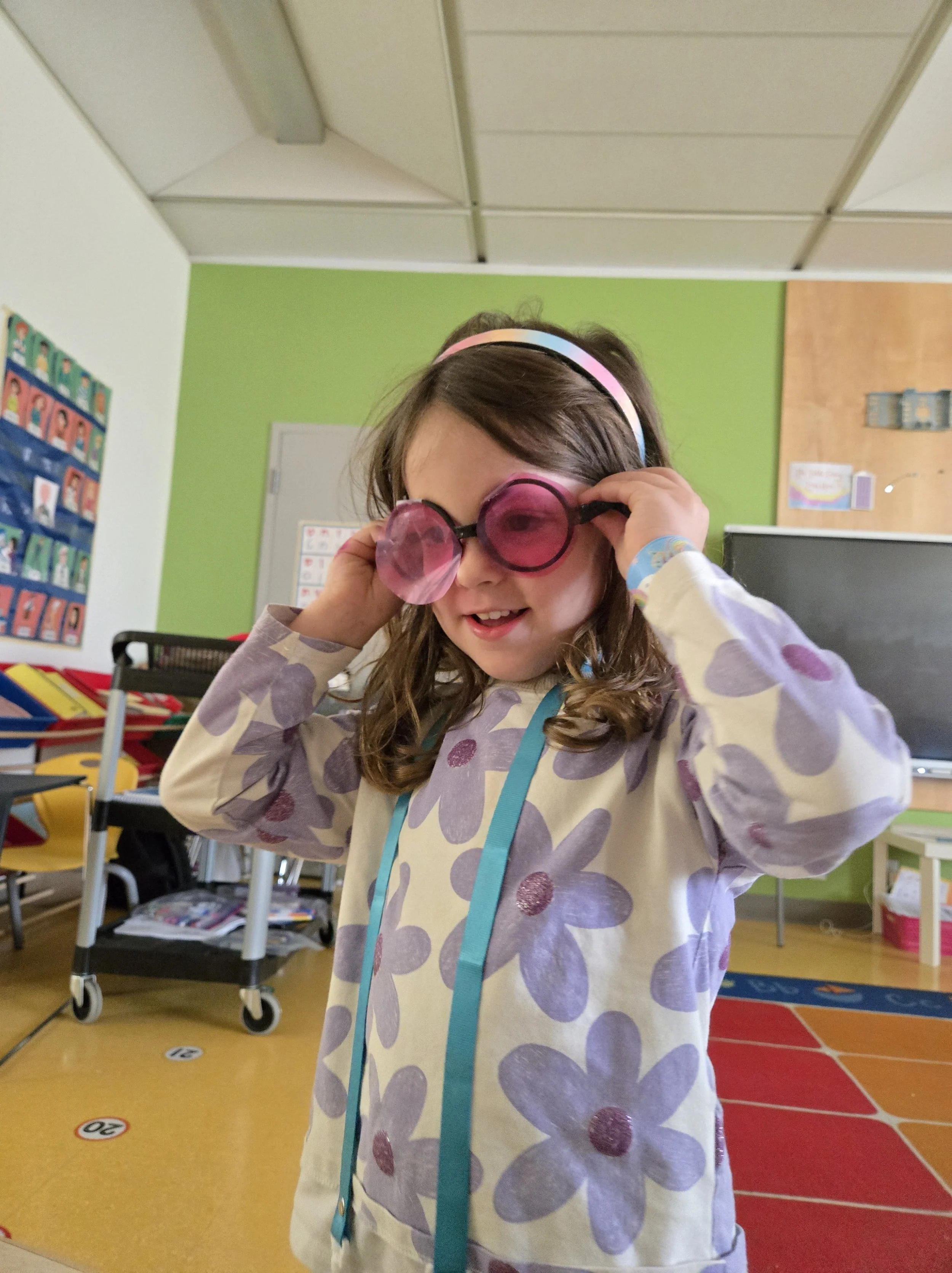 A young girl in a classroom trying on pink sunglasses and a headband, smiling and enjoying herself.