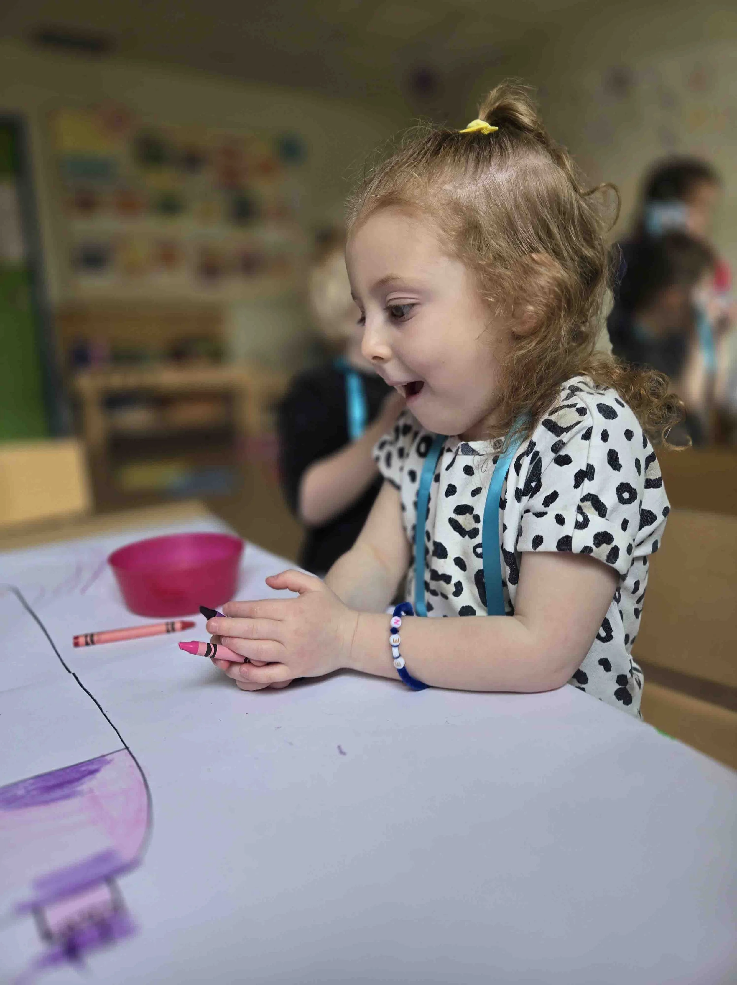 A young girl with curly brown hair in a ponytail, wearing a black and white leopard print shirt, is sitting at a table with a crayon in her hand. She appears excited and is looking at something on the table, which has a piece of paper and a pink bowl.