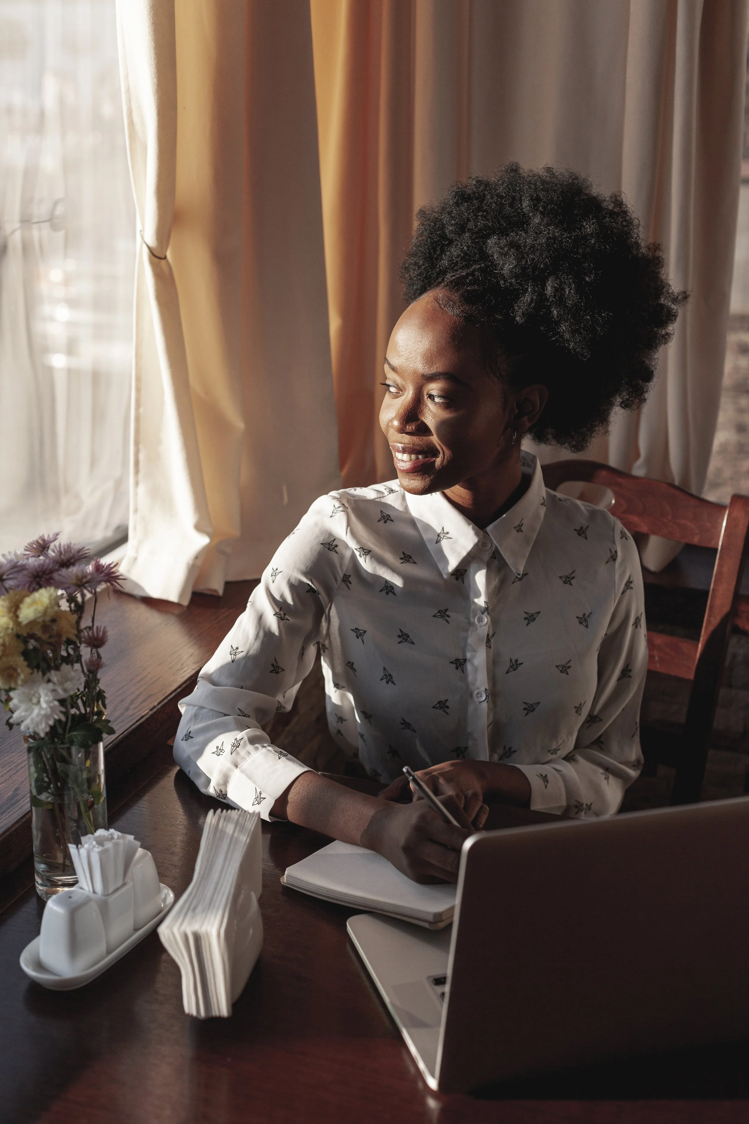 Woman sitting by a window writing in a notebook with a laptop open on the table in warm natural light.