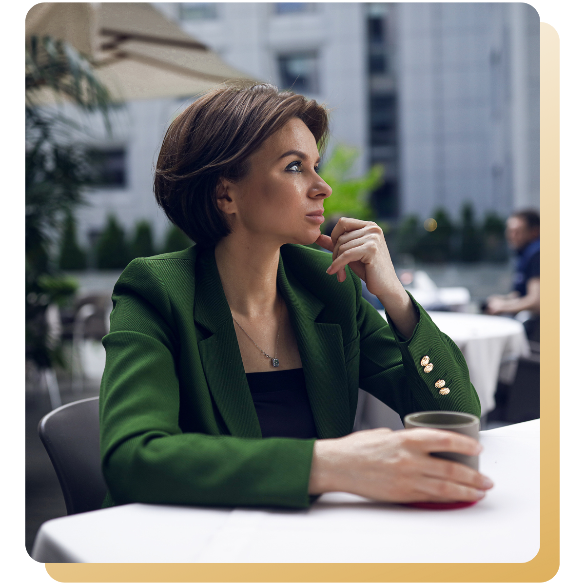 Professional woman in a green blazer sitting at an outdoor café, thoughtfully looking to the side while holding a cup.