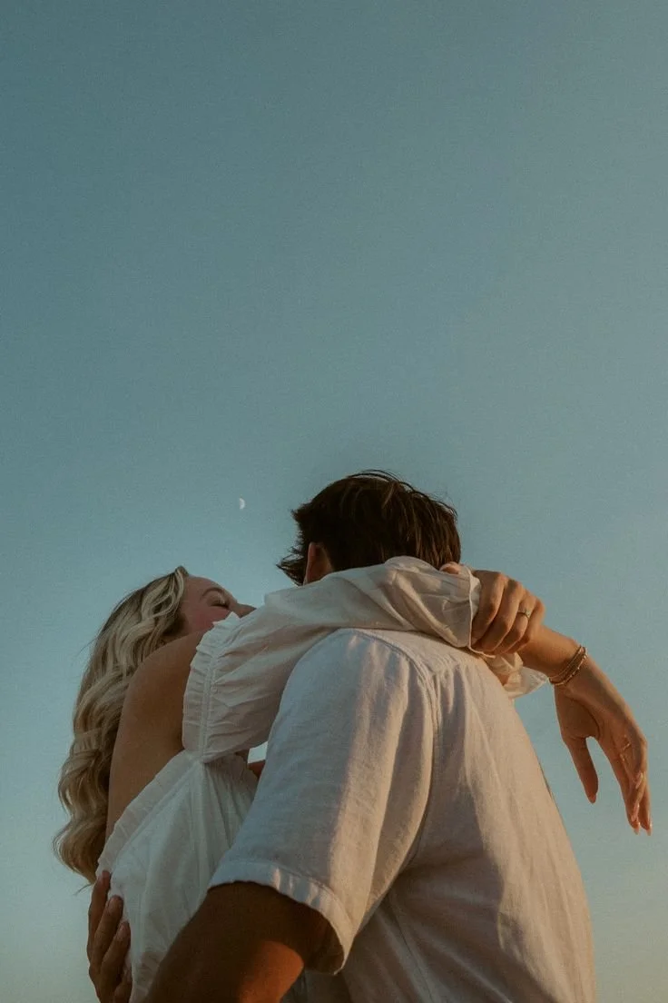 A couple embracing outdoors under a clear sky with a crescent moon visible, with the woman smiling and the man holding her close.