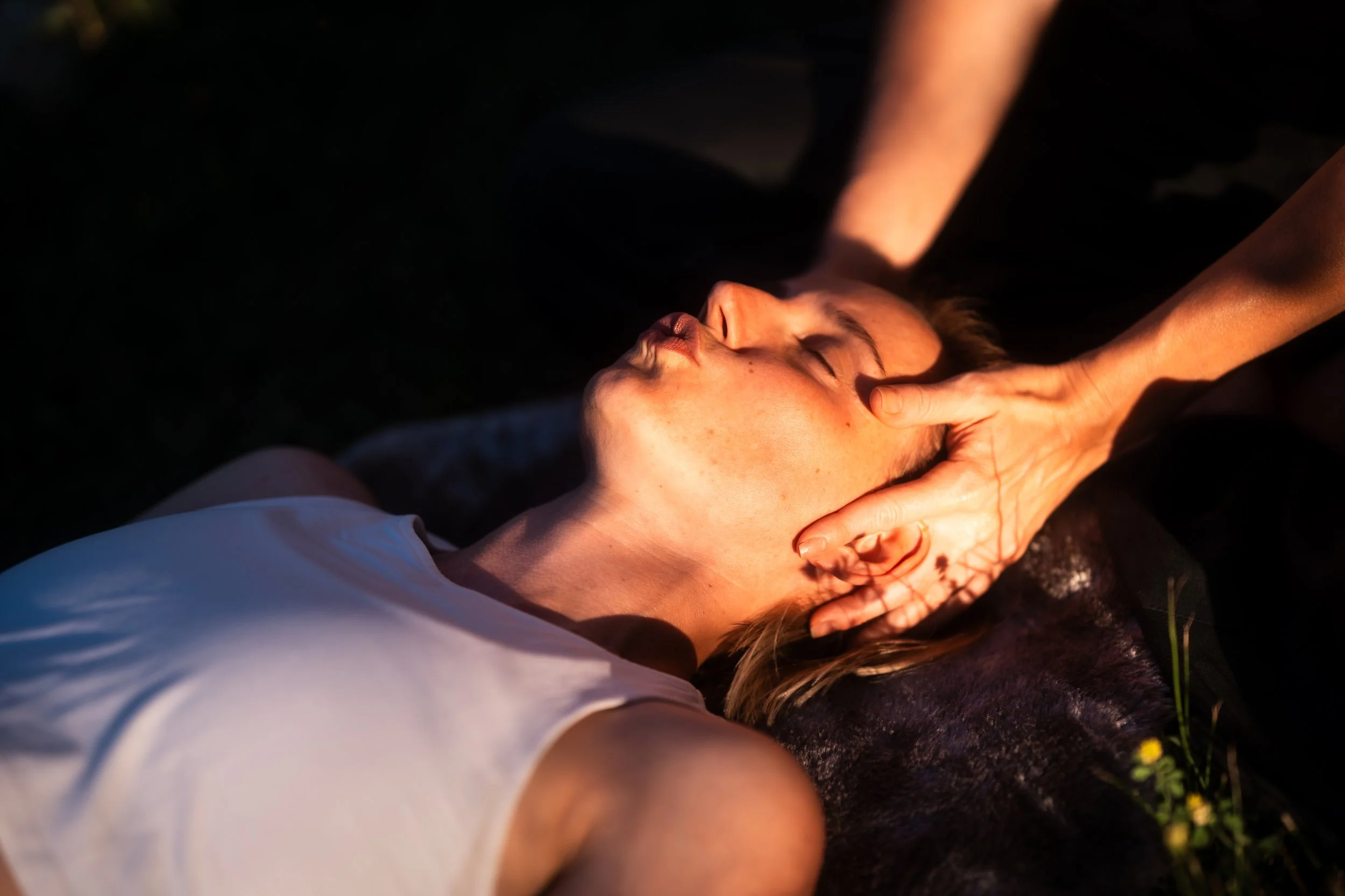 A woman lies on her back with eyes closed, receiving a head massage outdoors during sunset or sunrise, with sunlight illuminating her face.