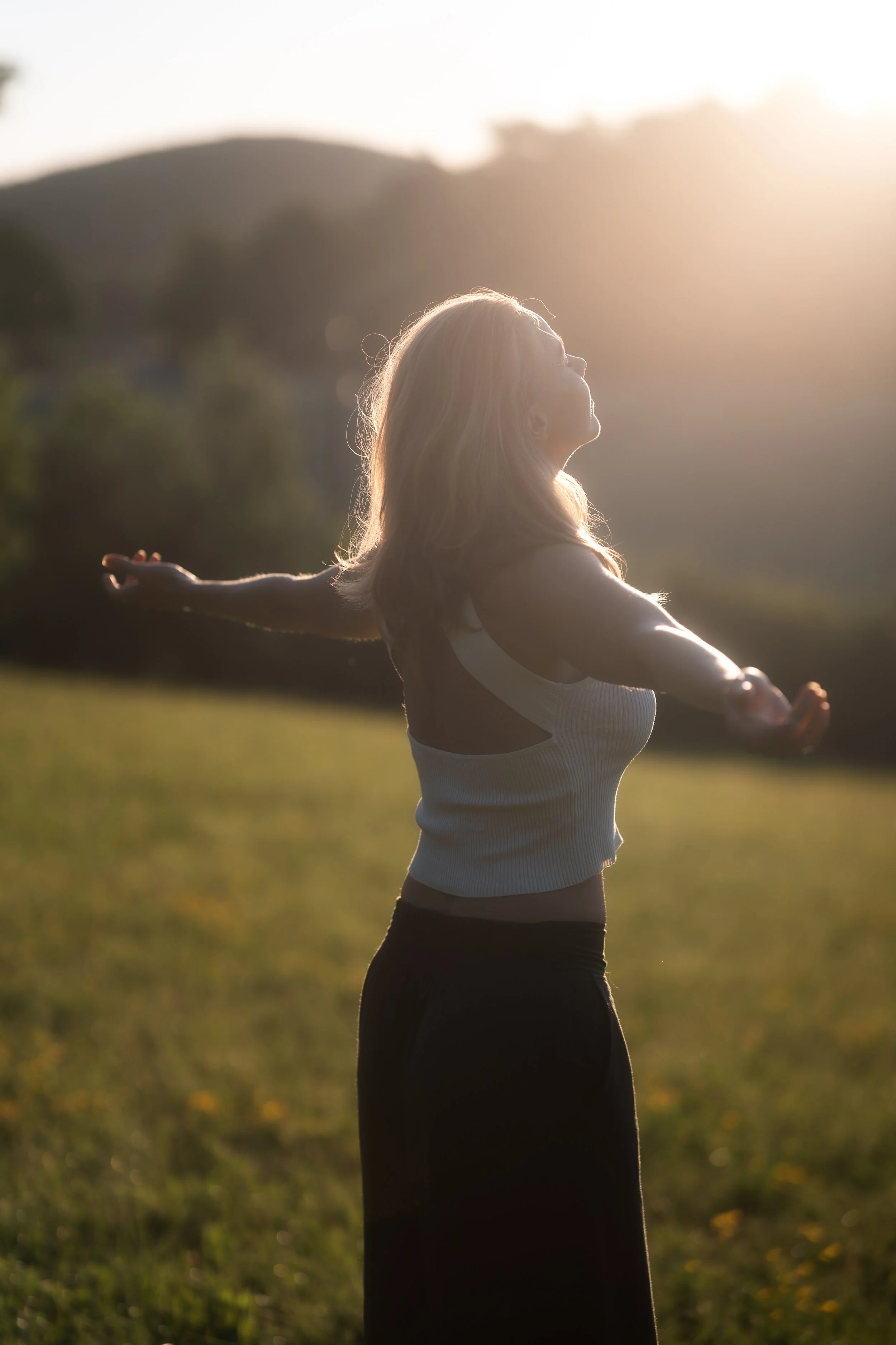 A woman in workout clothes standing outdoors with arms outstretched, enjoying sunlight and nature.
