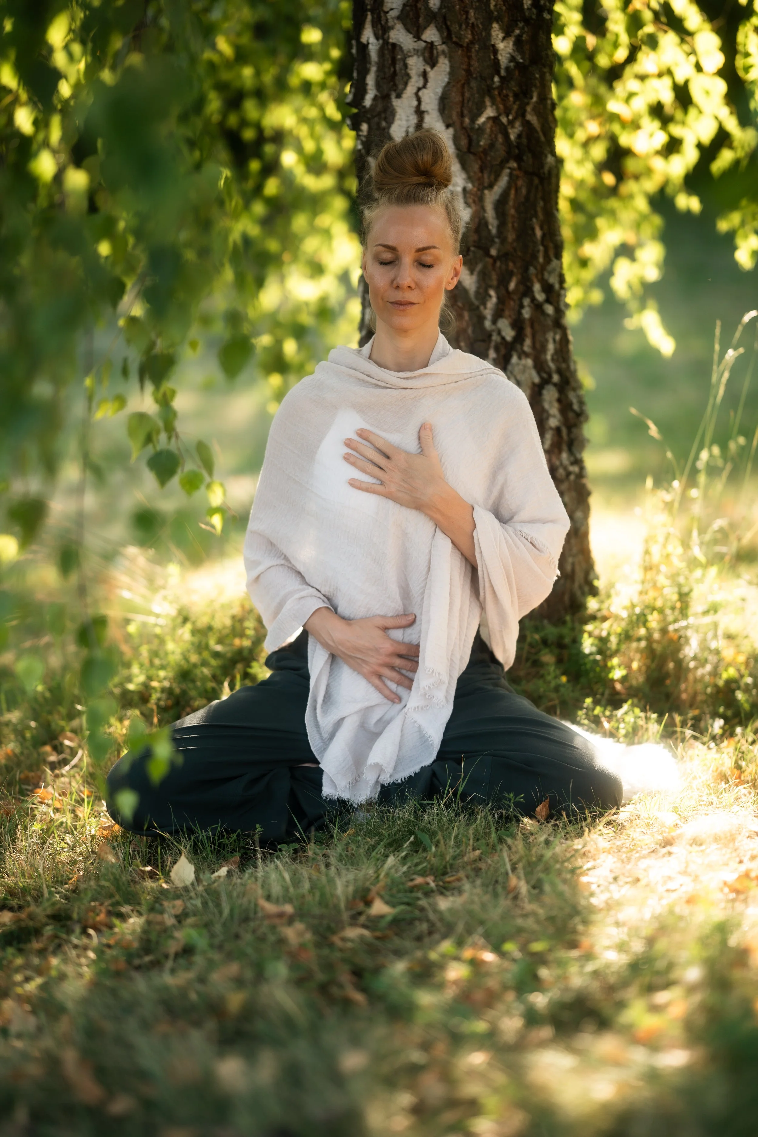A woman sitting cross-legged outdoors, holding her chest and abdomen with a peaceful expression, in a forested area with sunlight filtering through the trees.