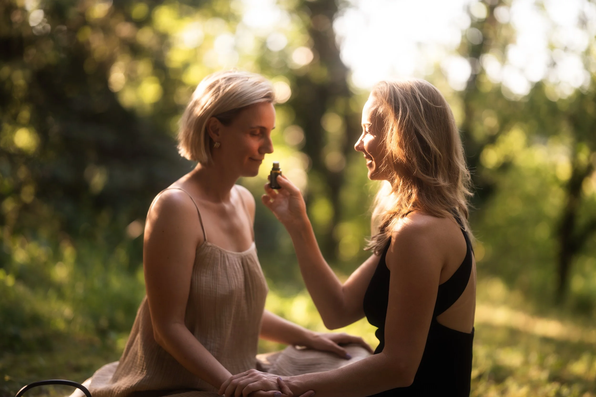 Two women sitting outdoors in a forest, one applying essential oil to the other's face, surrounded by sunlight and greenery.
