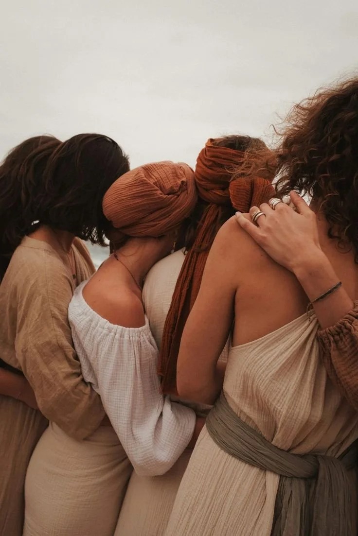 Four women standing close together, embracing and resting their heads on each other's shoulders, in a line against a plain white background, dressed in earthy-toned clothing.
