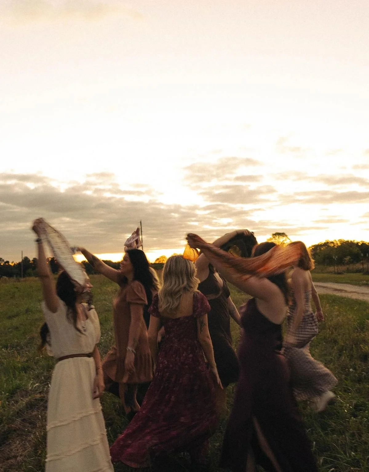 Group of women wearing bohemian dresses, dancing outdoors at sunset on a grassy field with a cloudy sky.