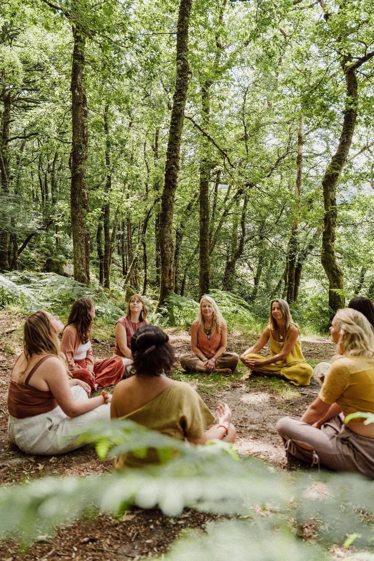 Group of women sitting in a circle on the forest ground, participating in a meditation or group session surrounded by tall green trees.