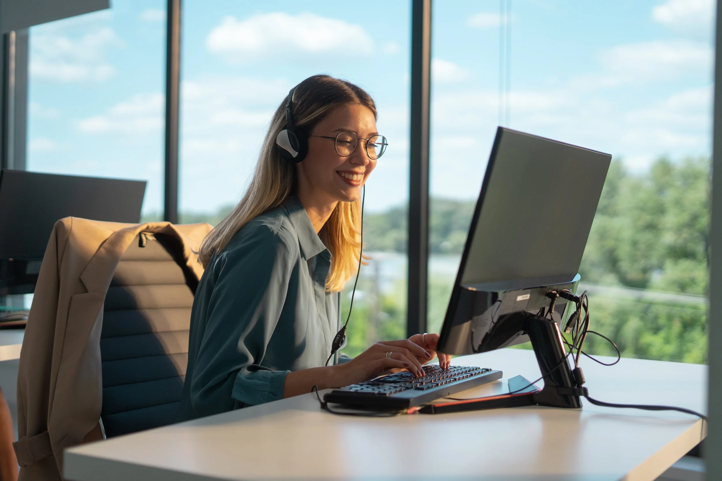 Una donna sorridente con cuffie al computer in un ufficio dall'aspetto moderno, con grandi finestre che mostrano un paesaggio verde e cielo blu.
