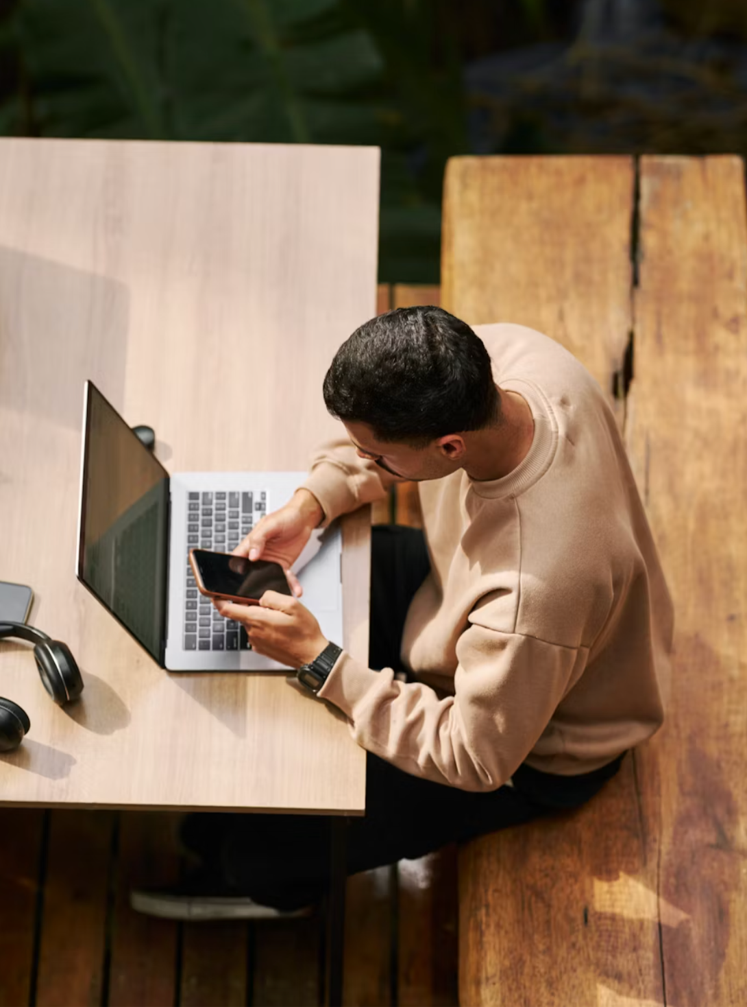 Un uomo seduto a un tavolo di legno che utilizza un computer portatile e uno smartphone, con cuffie nere e un orologio al polso, in un ambiente interno con sfondo di piante verdi.