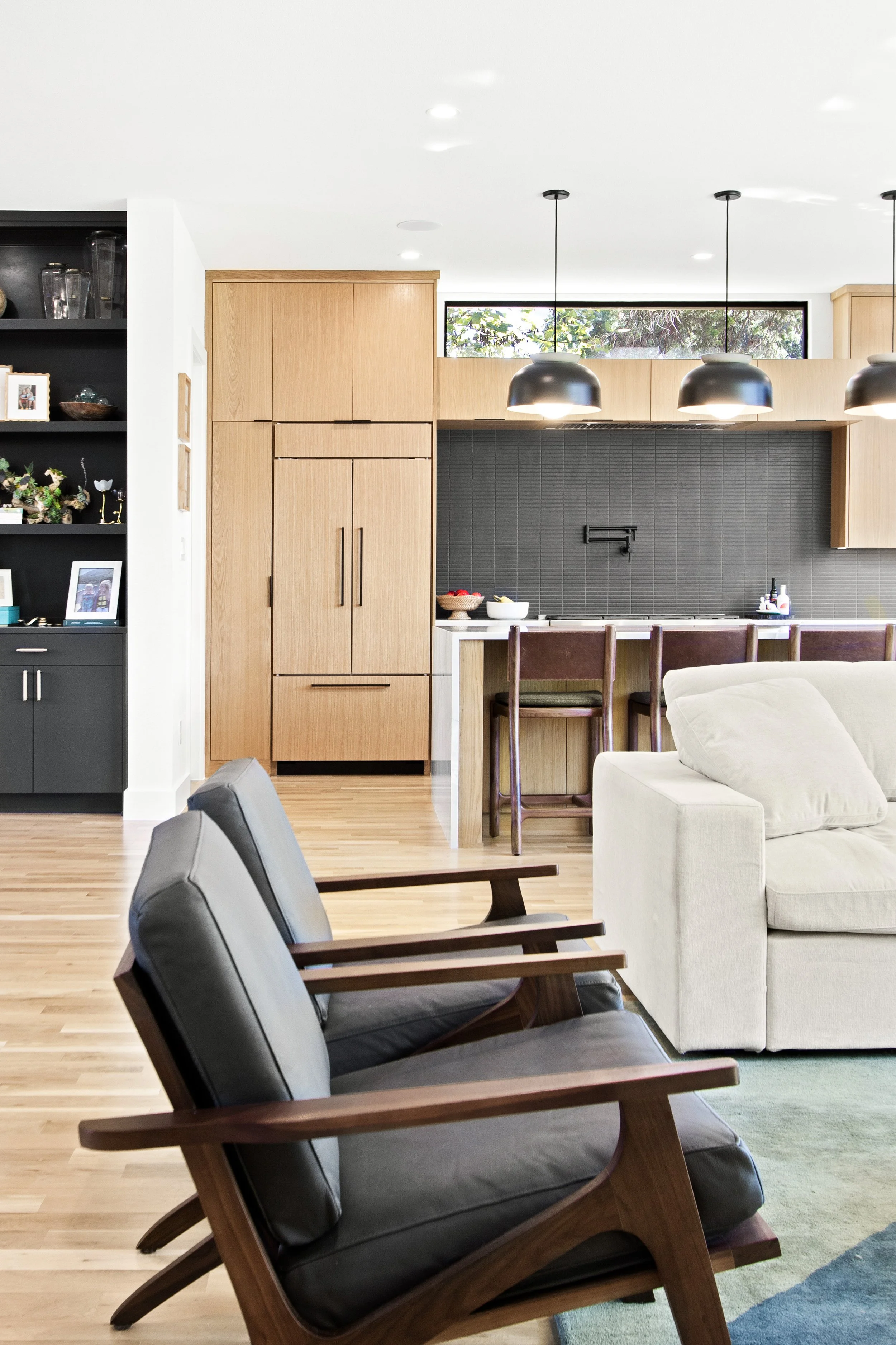 Modern living room with black armchairs, white sofa, black pendant lamps, wooden kitchen cabinets, black backsplash, and a breakfast bar with chairs.