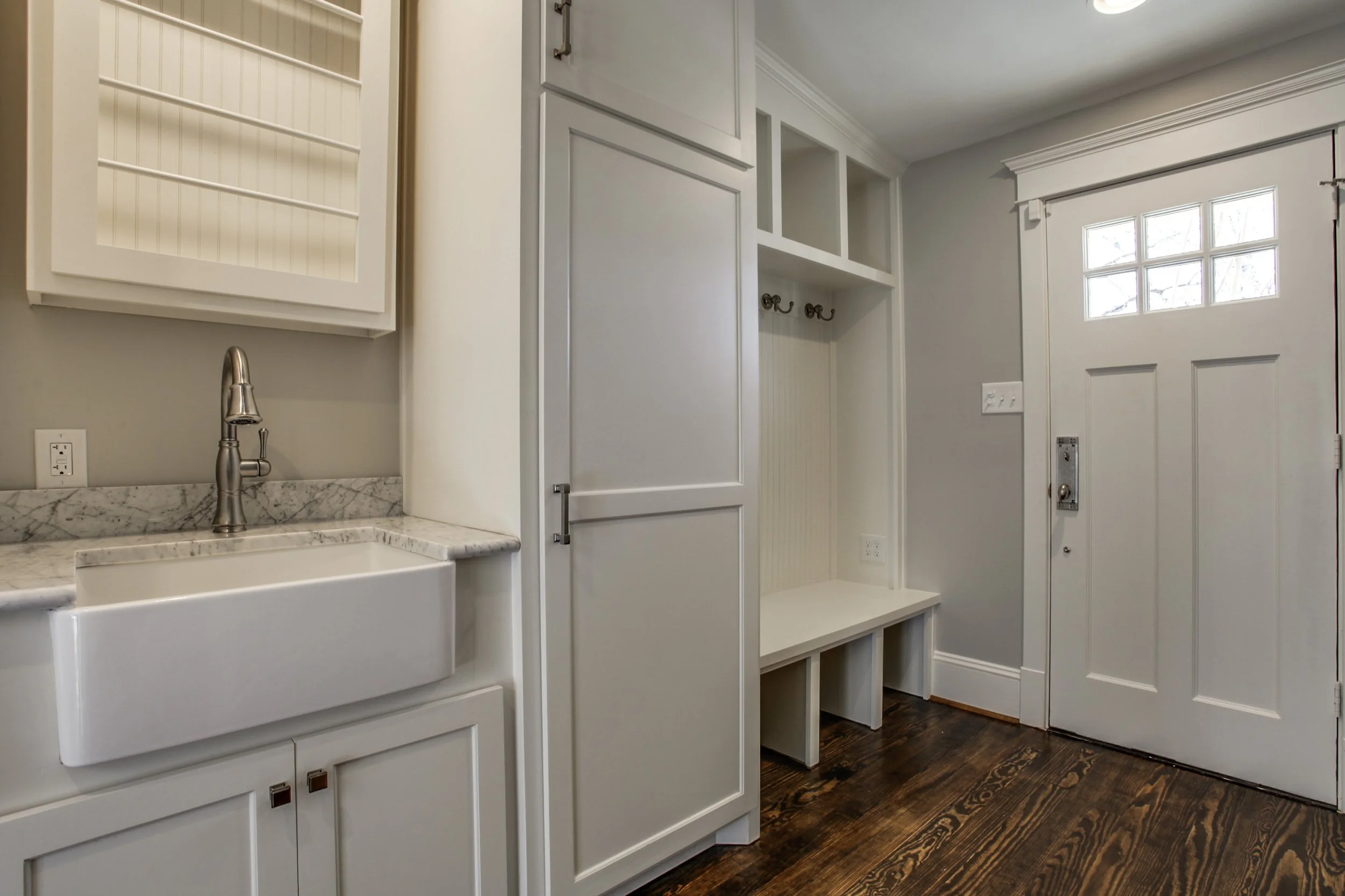Entryway with white cabinetry, a marble countertop, a white farmhouse sink, a wall-mounted cabinet, and a wooden bench with storage, next to a door with small window panes, on a dark wooden floor.