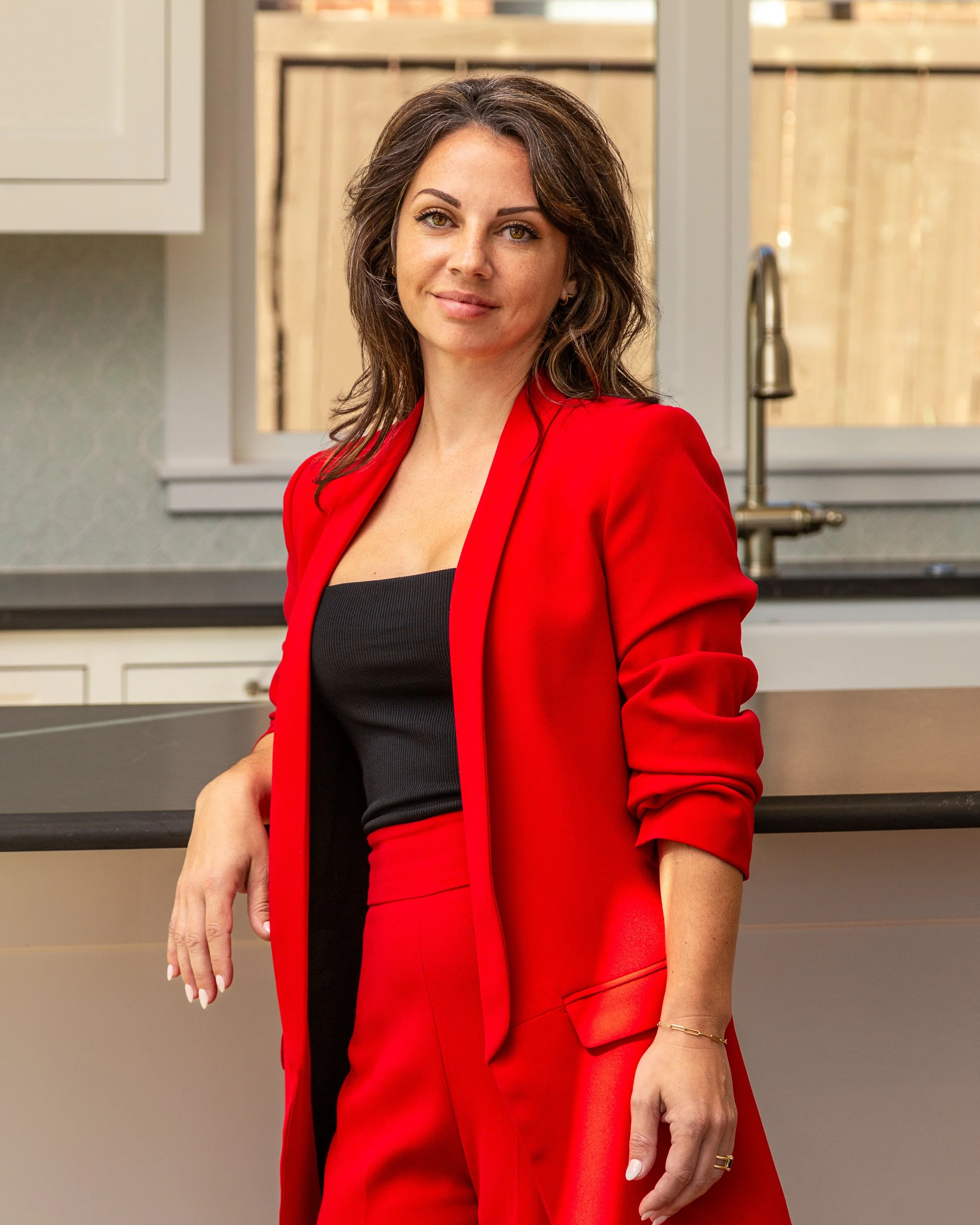 Woman with shoulder-length hair, wearing a red blazer and matching red pants, standing in a kitchen with a black top underneath, in front of a countertop and a window.