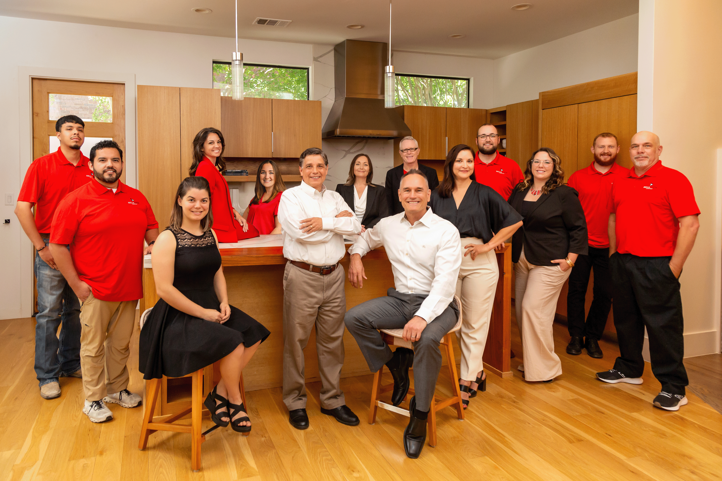 Group of 15 people in a modern kitchen, some wearing red shirts, posing for a photo. Two men are seated, others are standing behind them, smiling at the camera.