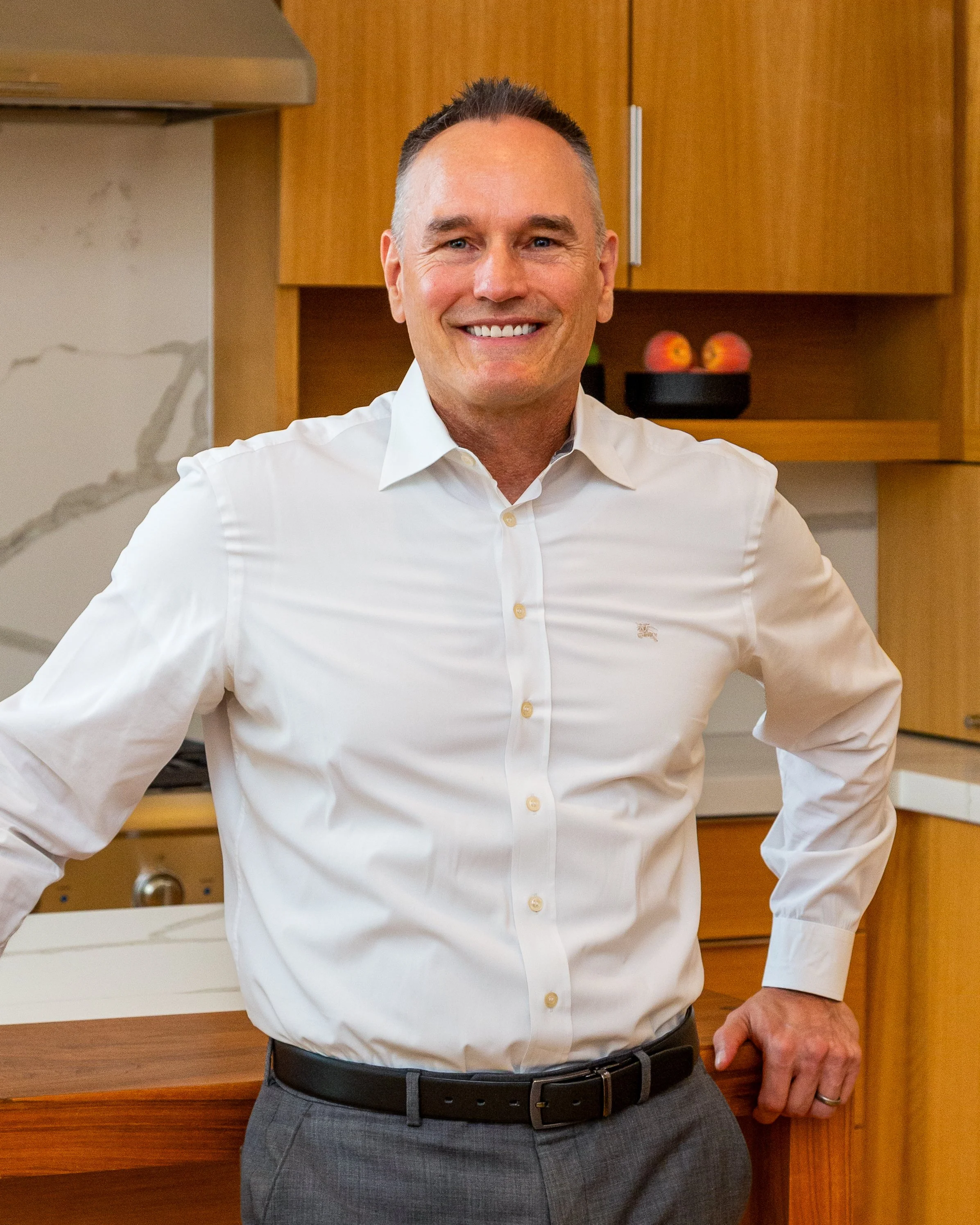 Smiling man in white dress shirt and gray pants standing in a kitchen with wooden cabinets and a white countertop.