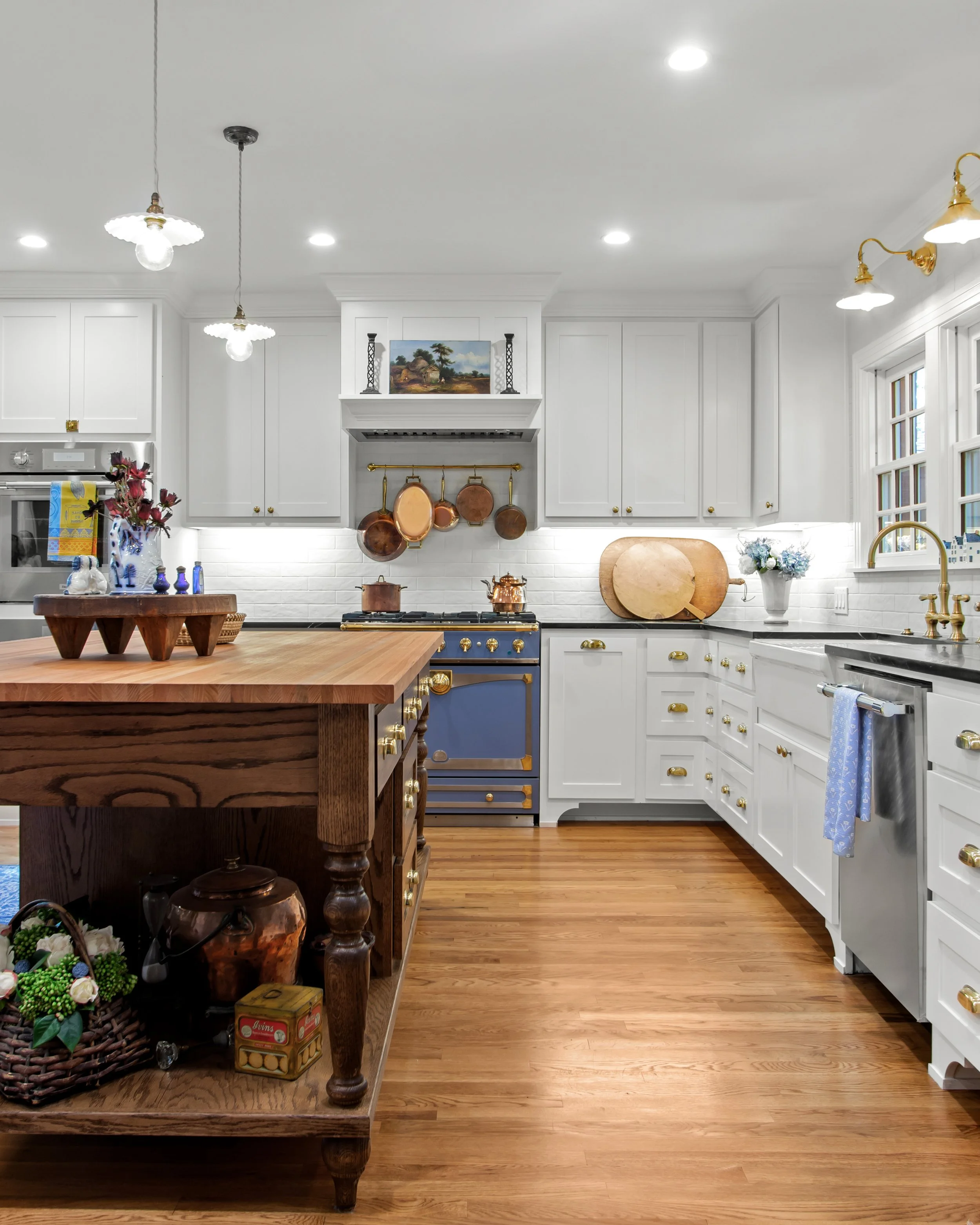 A bright kitchen with white cabinets, a wooden island, copper pots hanging above a blue stove, and hardwood flooring.