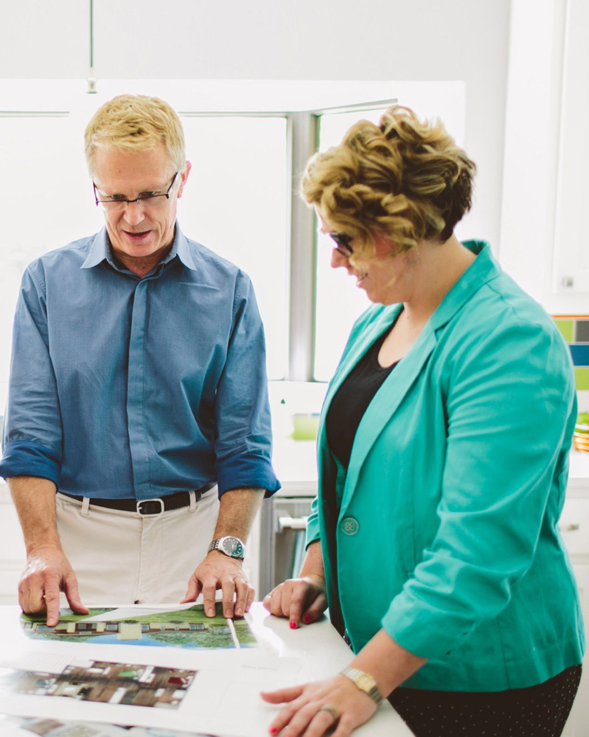 Two people, a man and a woman, are standing at a table looking at a large map or plan. The man is pointing at the map with his finger, and both are engaged in discussion. They are in a bright room with windows in the background.