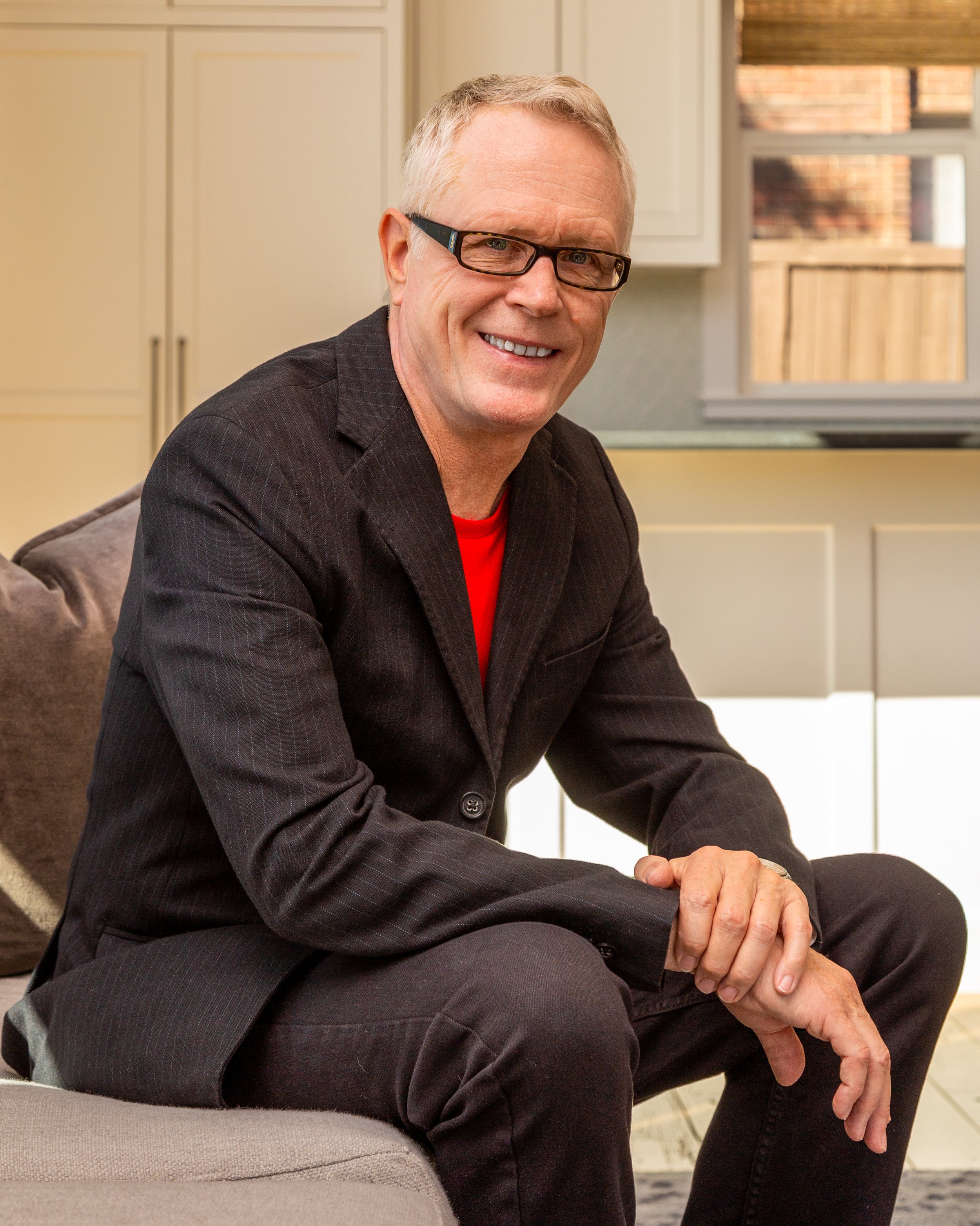A middle-aged man with glasses, smiling, sitting on a sofa in a modern living room.