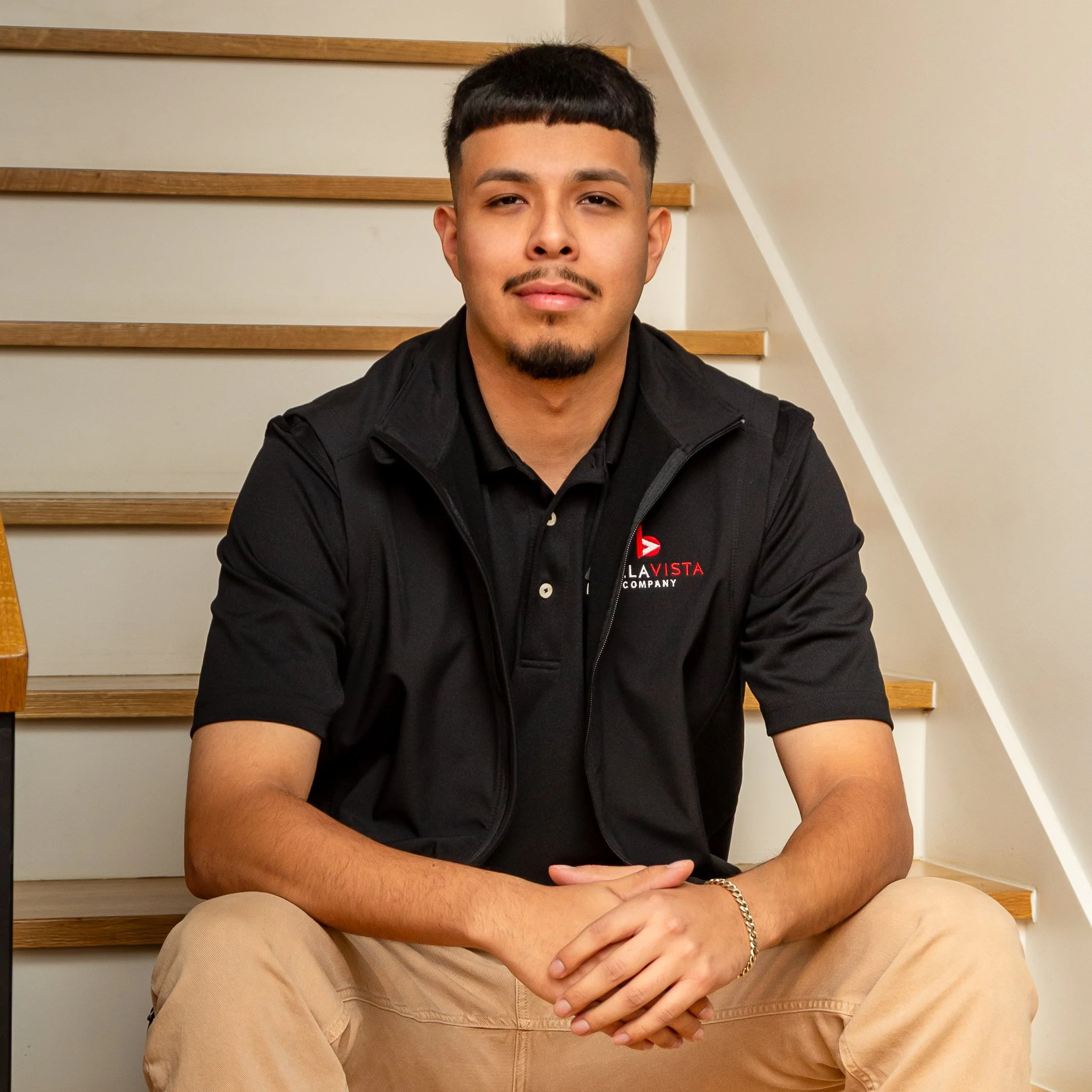 Man sitting on stairs, wearing a black polo and jacket, with beige pants, tattoos on his left arm, and a chain bracelet.