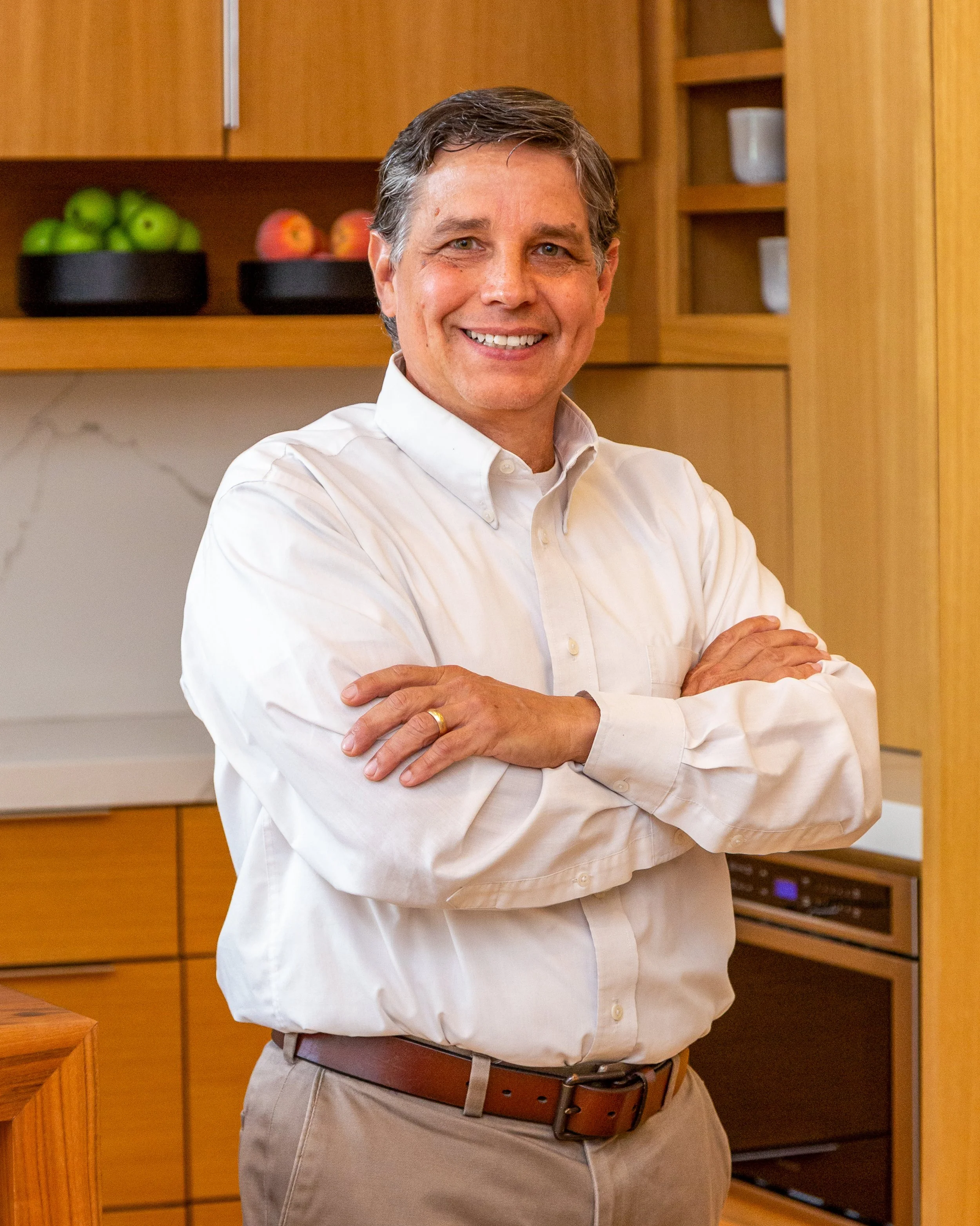 A man with short dark hair smiling and standing with his arms crossed in a kitchen with wooden cabinets and a bowl of green apples and red apples on a shelf behind him.