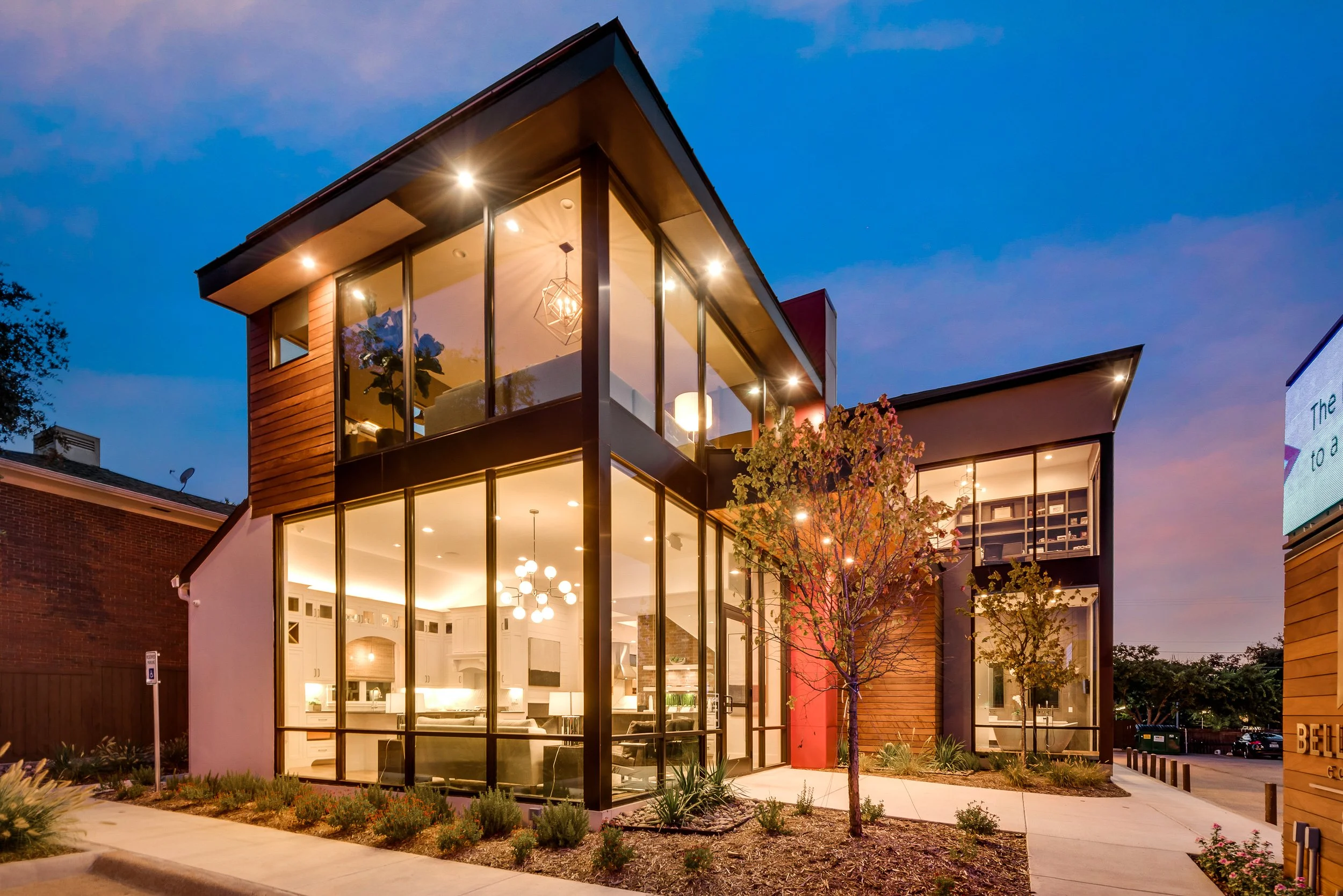 Modern house with large glass windows, illuminated interior, and landscaped front yard with small trees and plants during twilight.