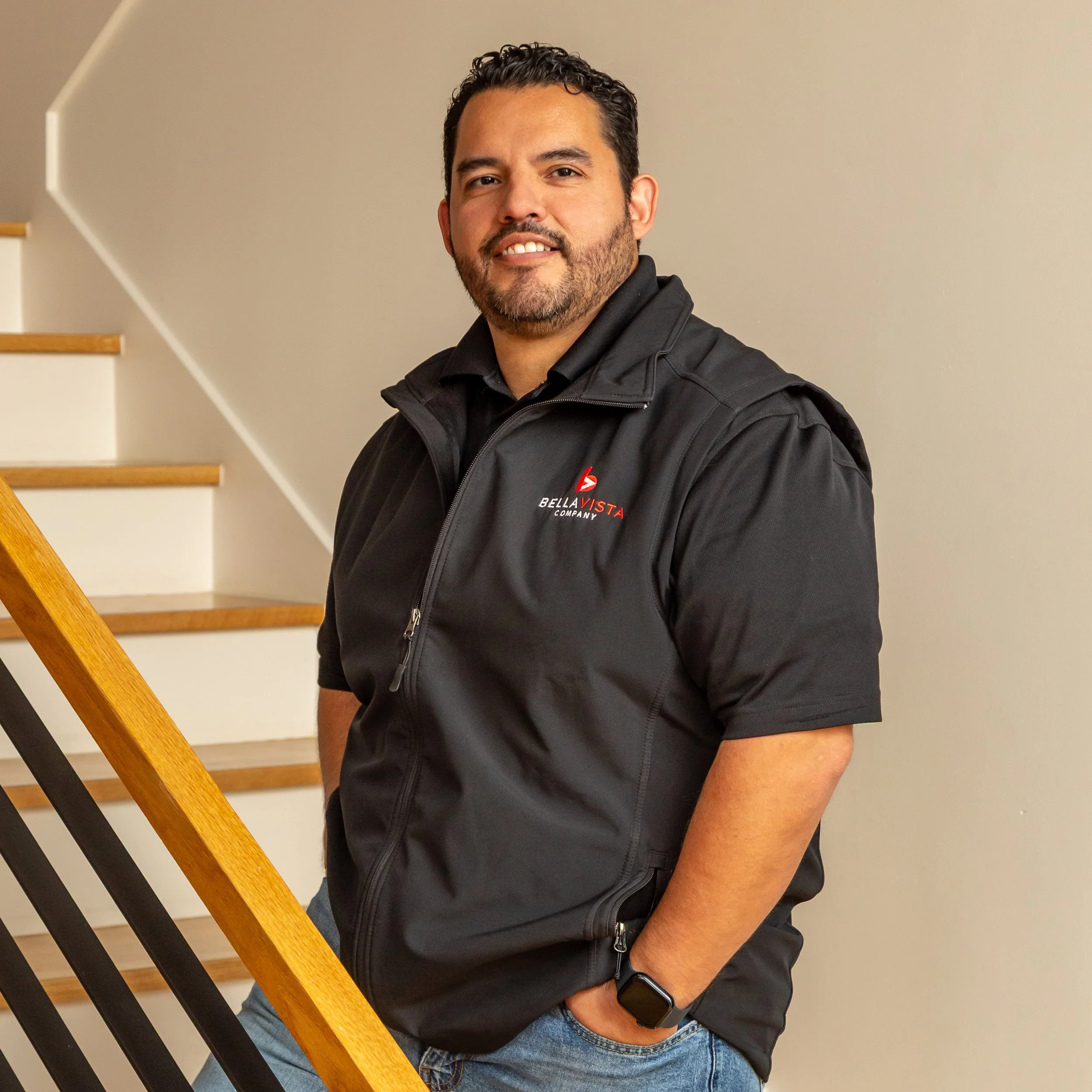 A man with dark hair and a beard standing indoors on a staircase, wearing a black vest with a logo that says Bella Vista Company, and a smartwatch on his left wrist.