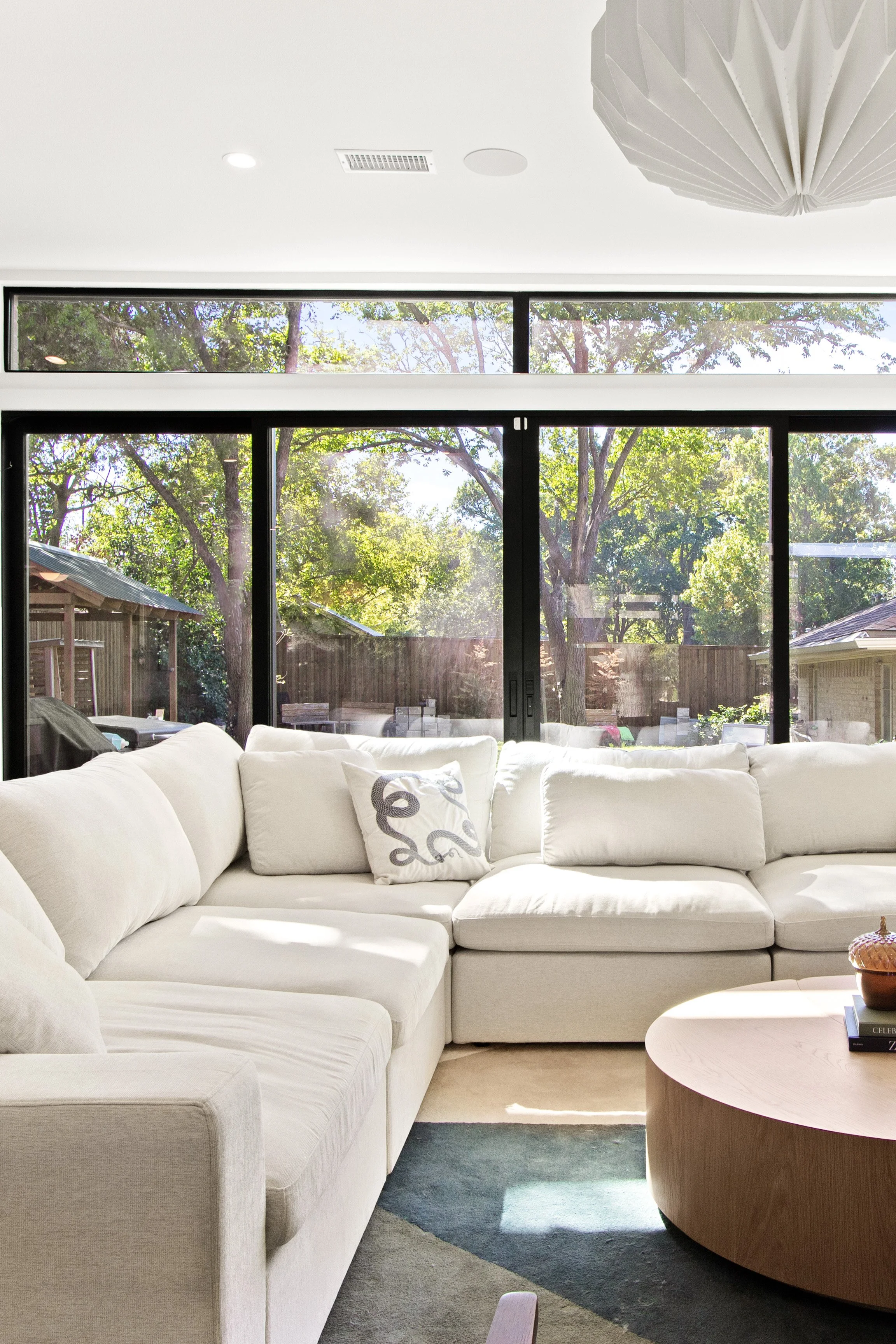 Modern living room with a large white sectional sofa, a decorative pillow with an octopus design, a round wooden coffee table, and large sliding glass doors leading to an outdoor backyard with trees and a wooden fence.