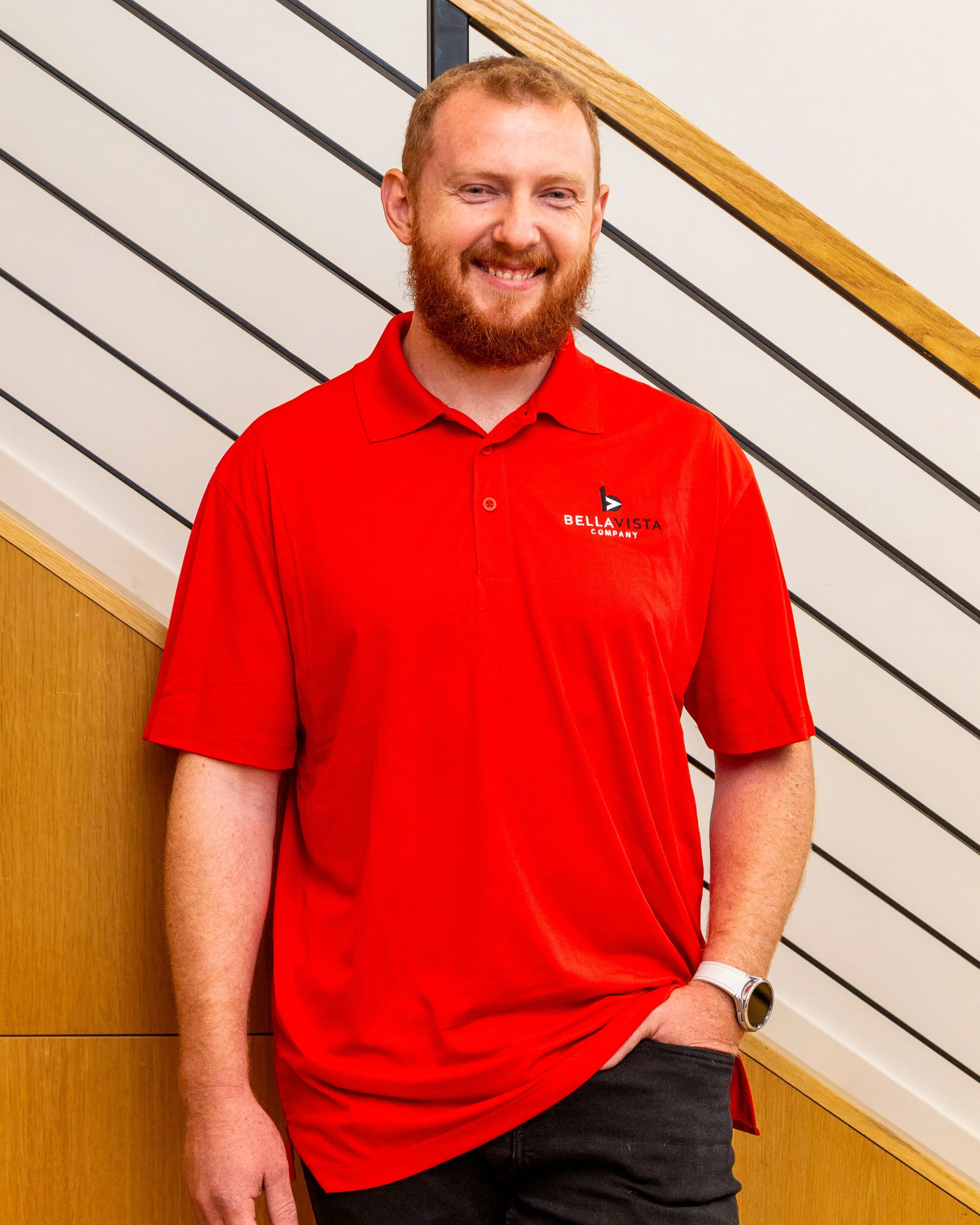 A man with red hair and a beard wearing a red polo shirt and a watch, standing against a wood-paneled wall and a staircase with black railings.