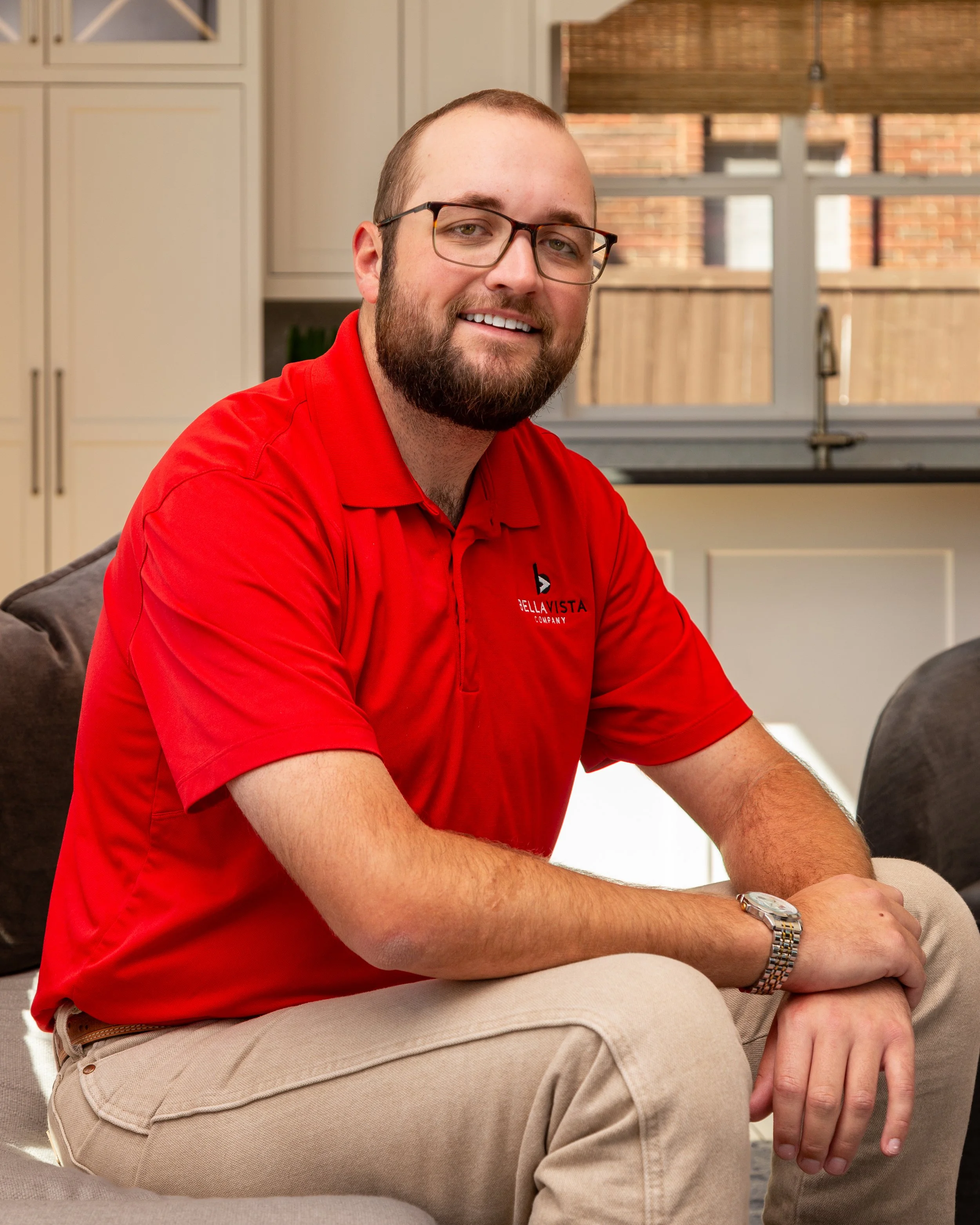 A man with glasses and a beard wearing a red polo shirt and khaki pants sitting on a gray couch in a bright kitchen space.