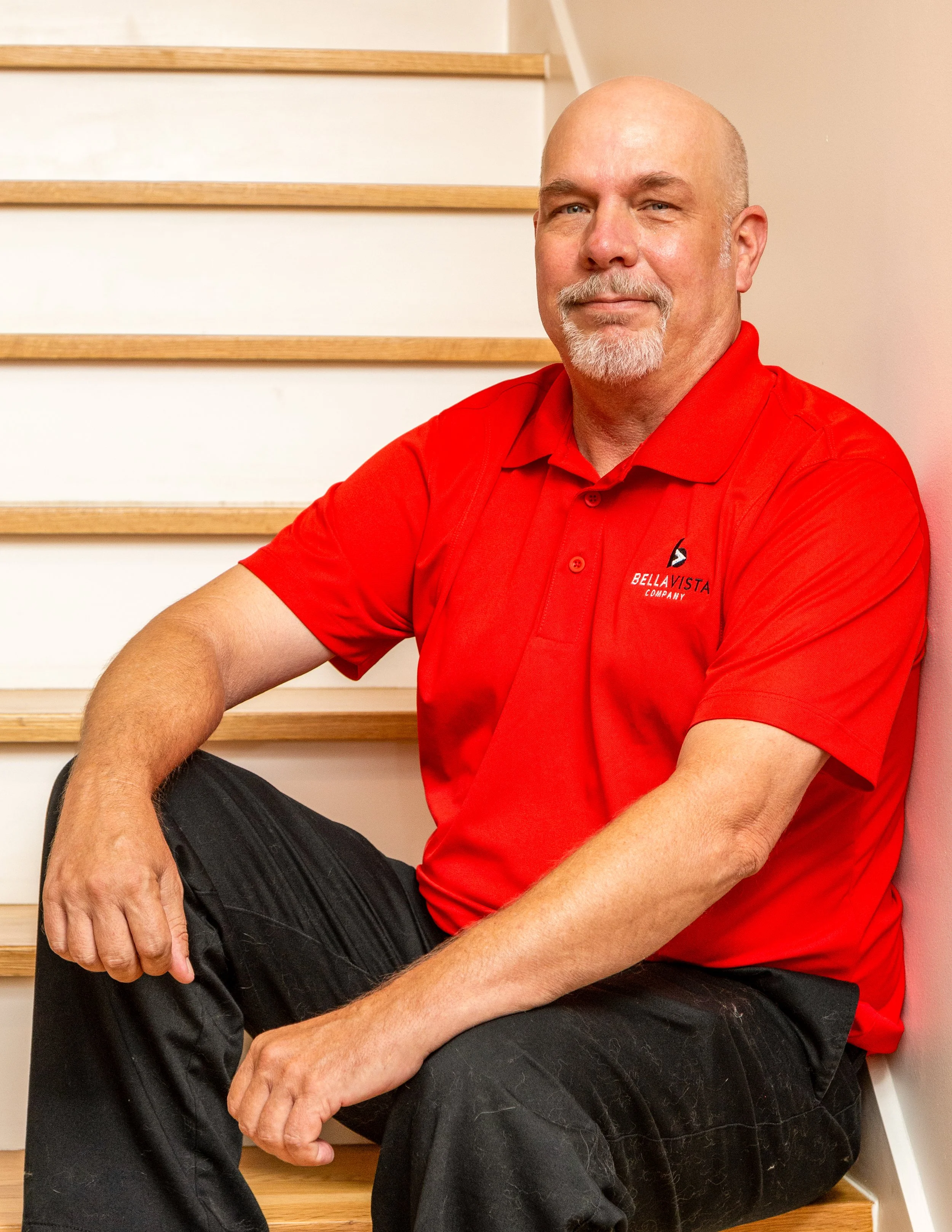 A bald man with a white beard wearing a red polo shirt with a logo, sitting on stairs and smiling.