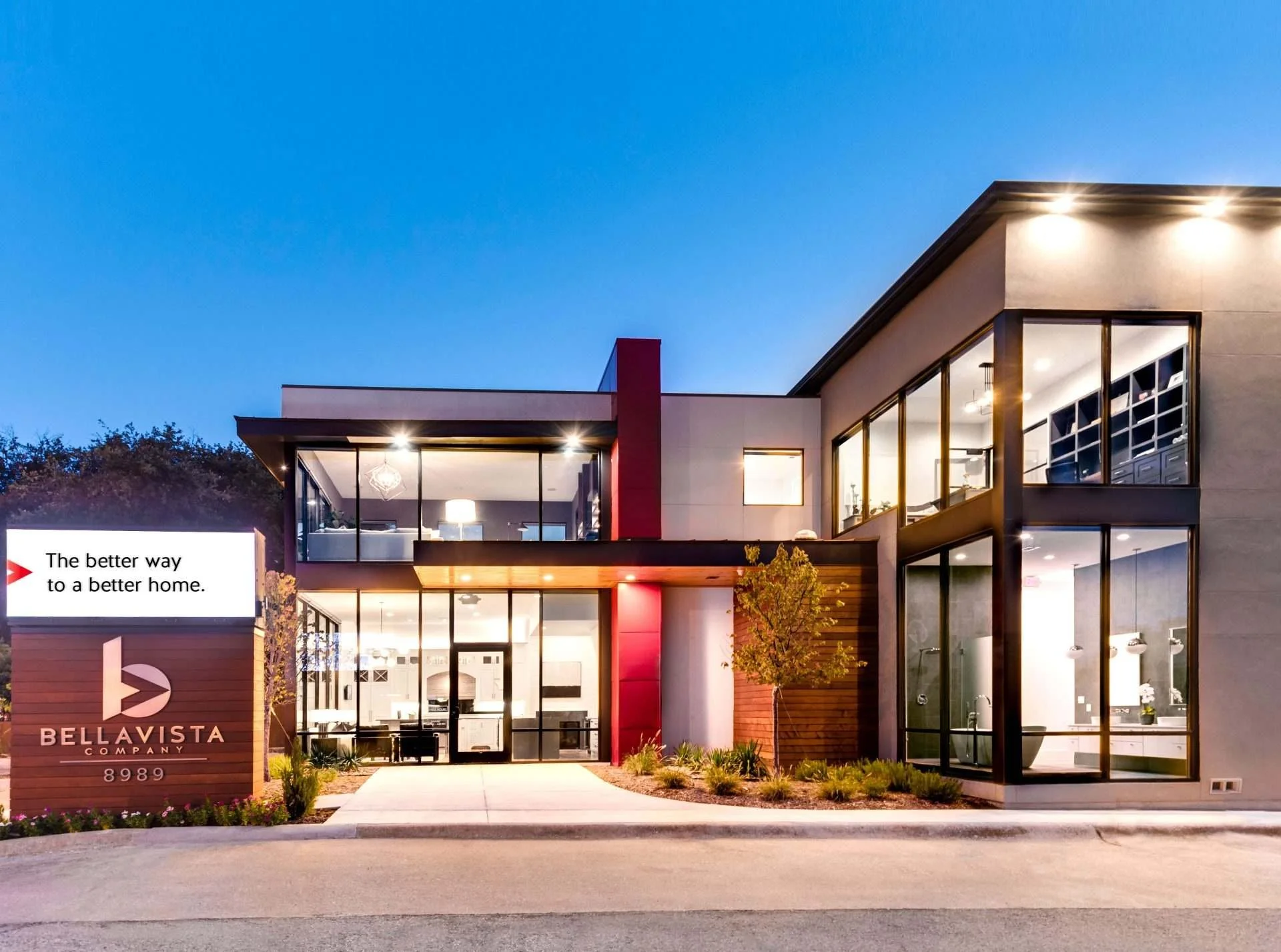 Modern two-story building with large glass windows, outdoor lighting at dusk, landscaped surroundings, sign that reads 'Bellavista Company' with address 8989, and a tagline 'The better way to a better home.'