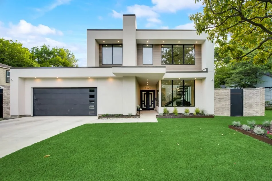 Modern two-story white house with large windows, a black garage door, and a manicured green lawn, set against a blue sky and trees.