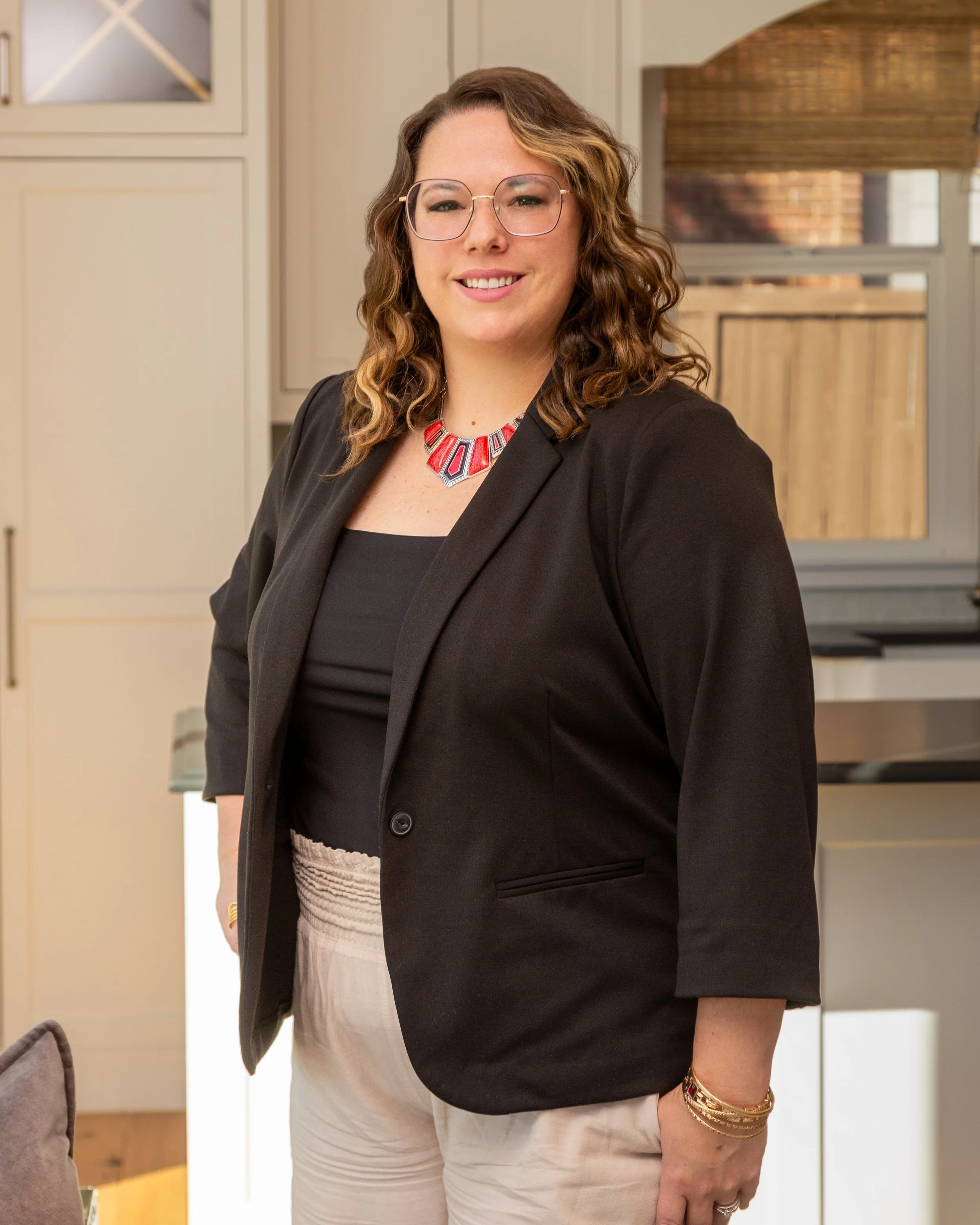 Woman with curly hair, glasses, dressed in a black blazer and white pants, standing in a bright kitchen.