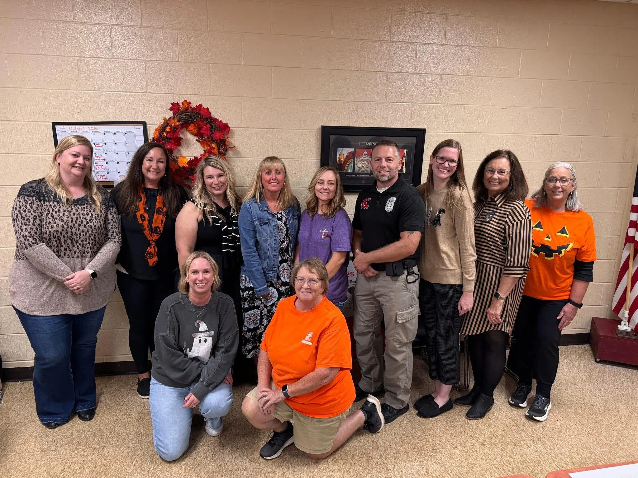 Group of fourteen people, including men and women, standing and kneeling in a room decorated with a seasonal wreath and pumpkins, many of whom are smiling for the camera. The room has a beige brick wall with a framed picture and a digital calendar visible in the background.