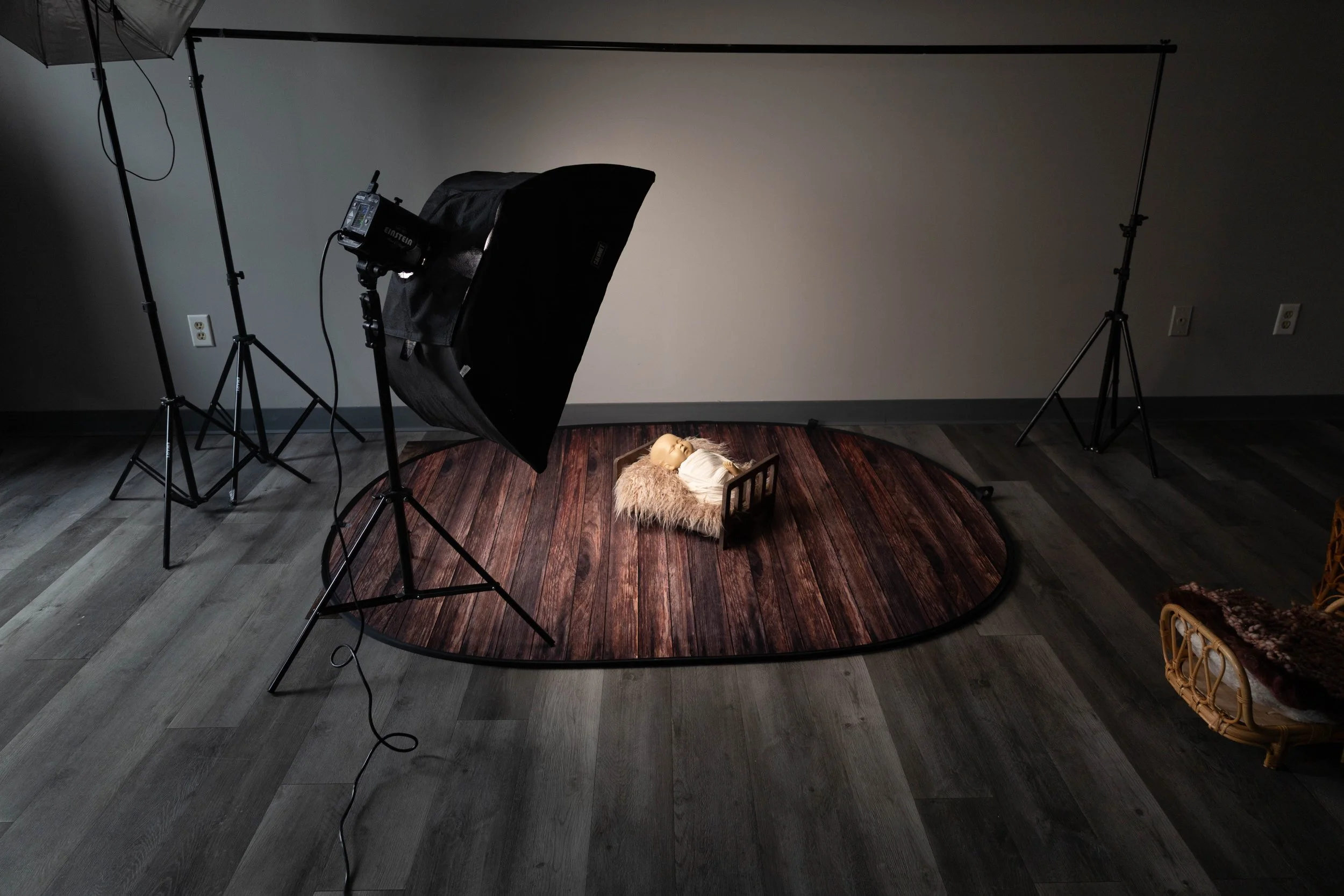 A photography setup with studio lights focused on a doll lying in a crib on a round wooden platform, all inside a room with wooden flooring.