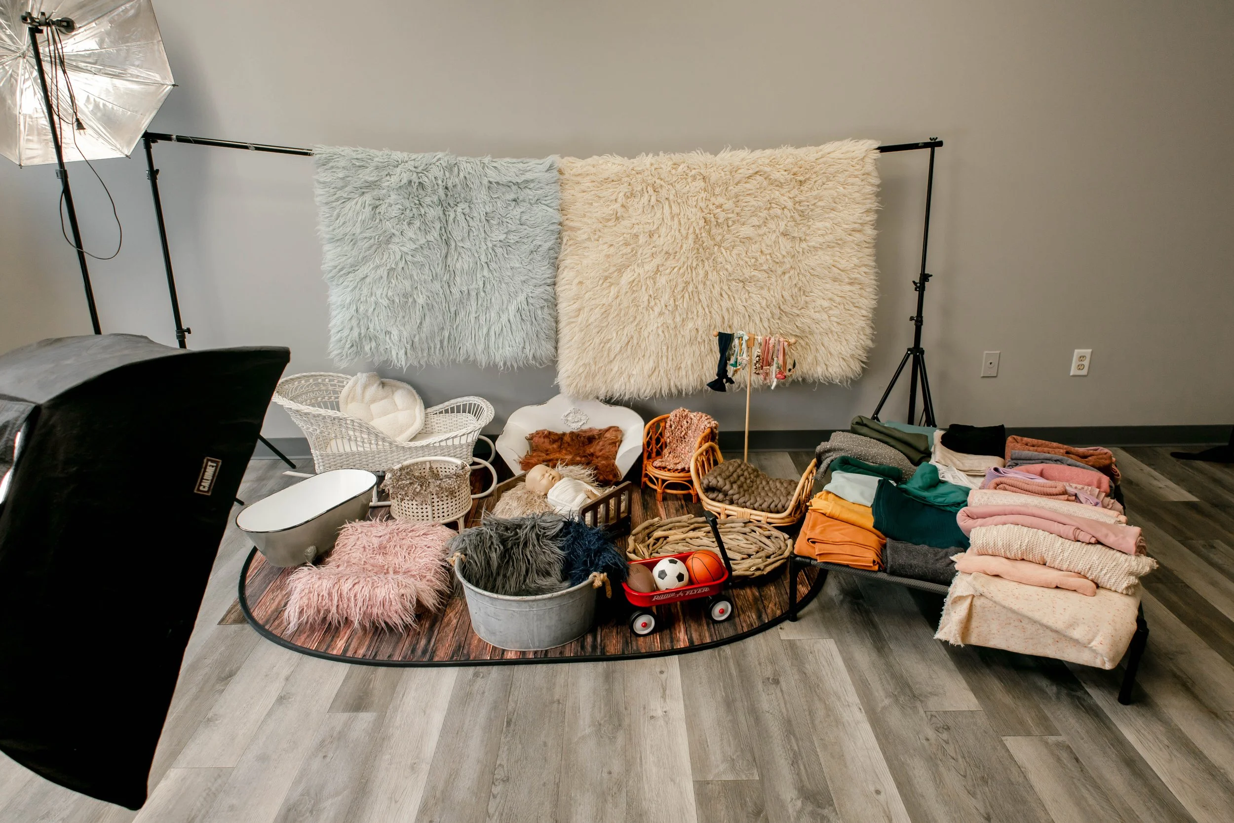 Photography setup with two large fluffy rugs hanging on a backdrop, various cozy baskets and props, clothing, and toys arranged on the floor, with studio lighting and equipment visible.