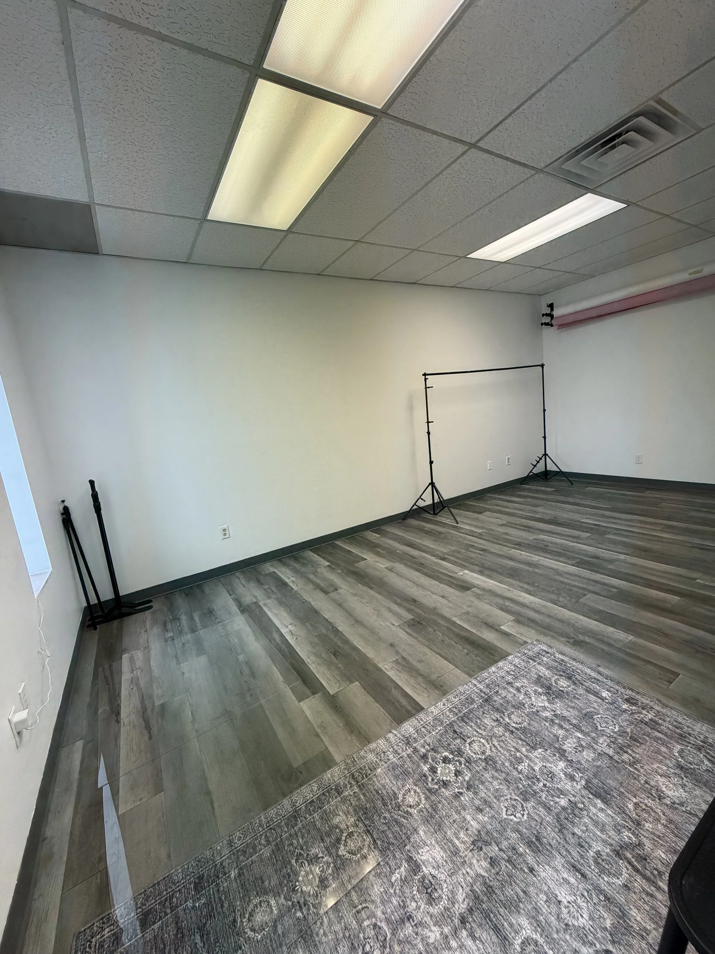Empty room with gray flooring, white walls, ceiling tiles with fluorescent lights, and photography equipment including a backdrop stand and light reflectors.