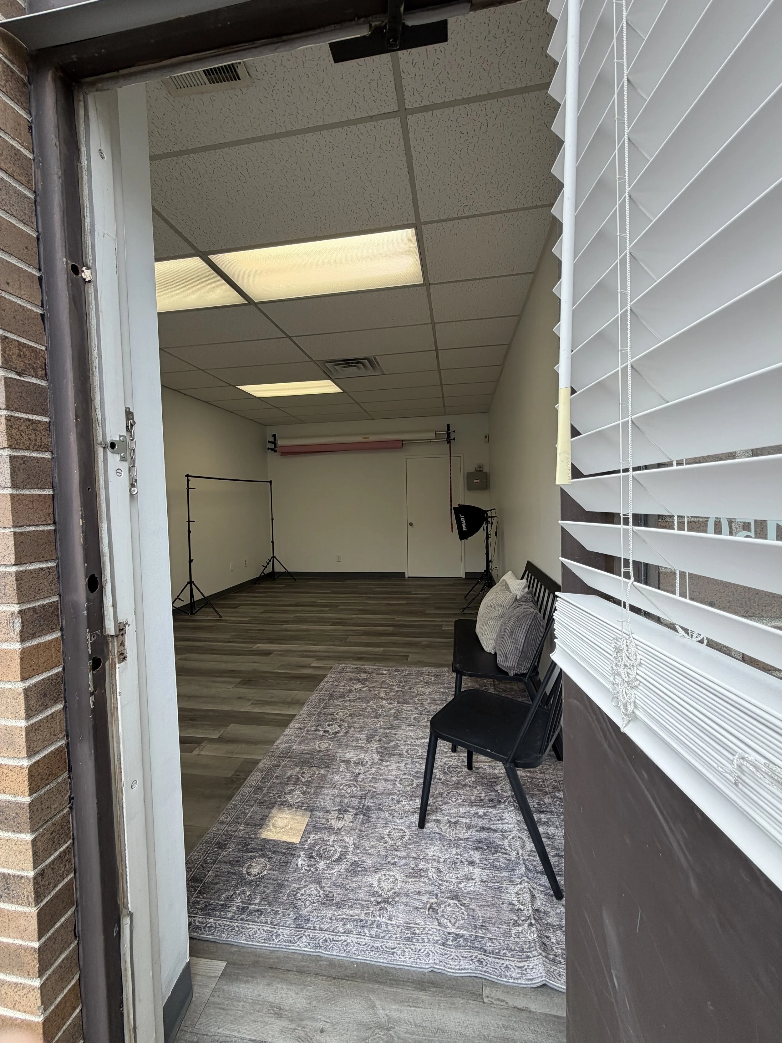 View from outside into a small room with a patterned rug on a wood floor, black chairs with pillows, and studio lighting equipment against a plain white wall, seen through an open door with white blinds.