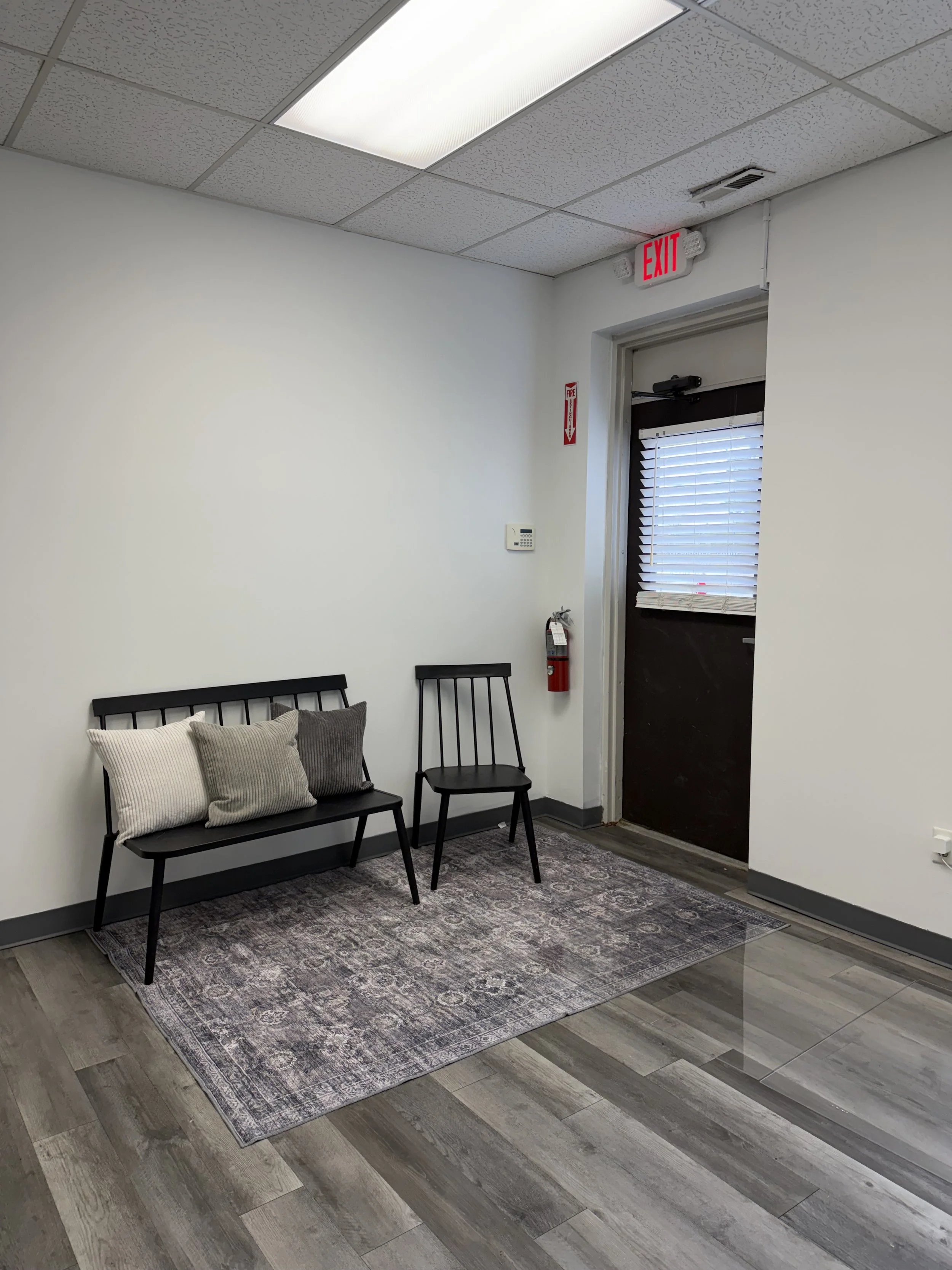 Waiting area with a black bench and single black chair, three pillows on the bench, a patterned rug, a fire extinguisher, and an exit door with a window with blinds.