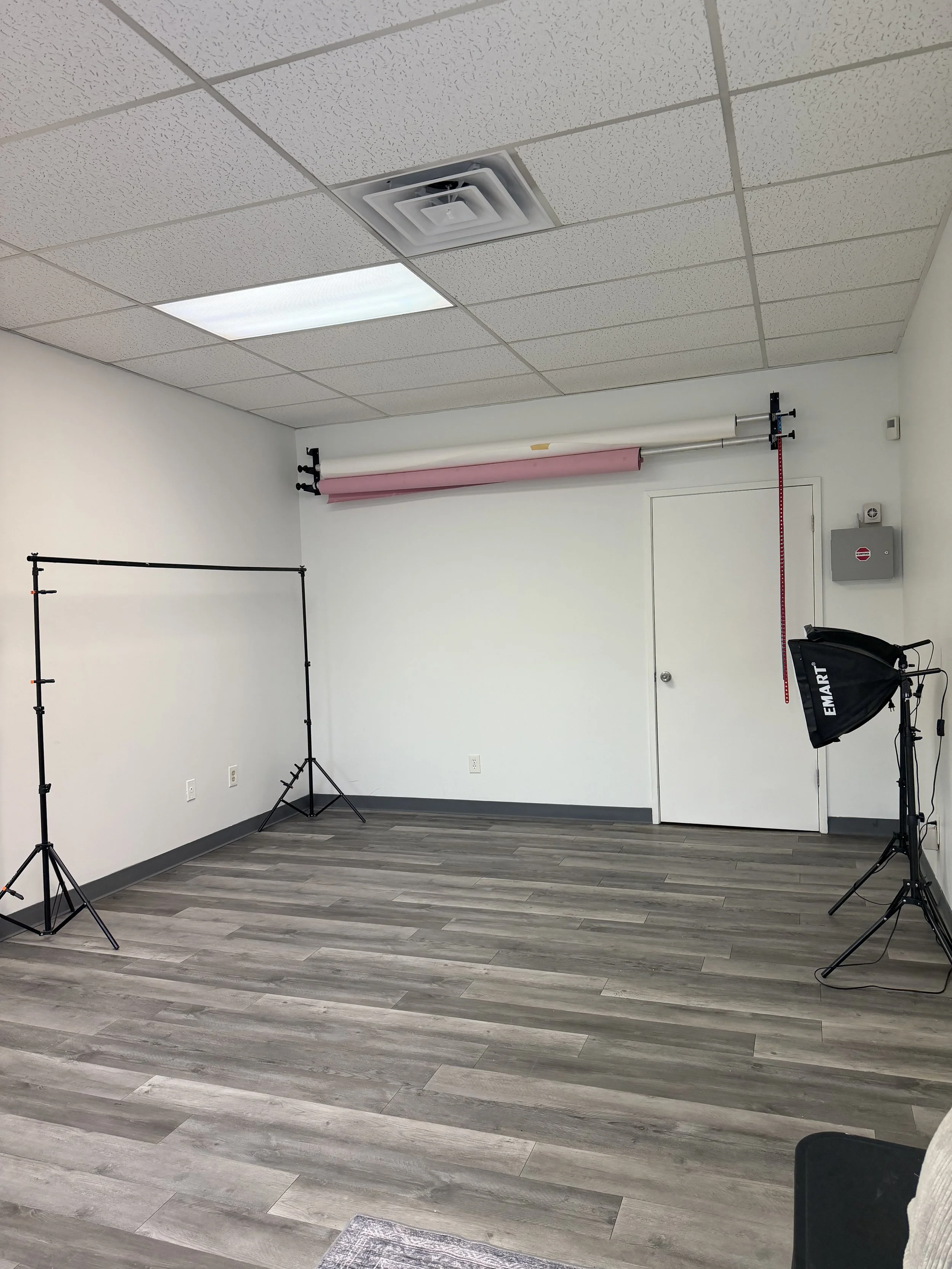 Empty photography studio with white walls, gray wooden floor, lighting equipment, and a backdrops roller at the back.